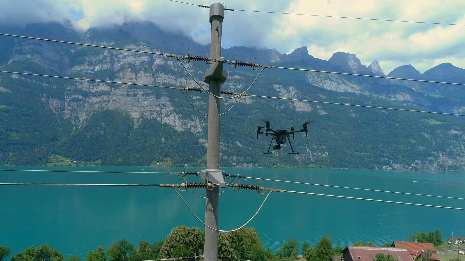 A drone flies near power lines with a lake and mountains in the background. The landscape includes a turquoise lake, green hills, and rugged cliffs under a partly cloudy sky.