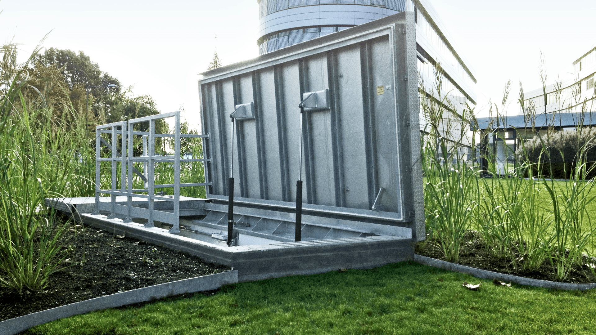 Ein großes Hochwasserschutzgitter aus Metall ist auf einer Wiese neben einem modernen Gebäude installiert, die teilweise offen ist und in deren Nähe hohe Gräser wachsen.