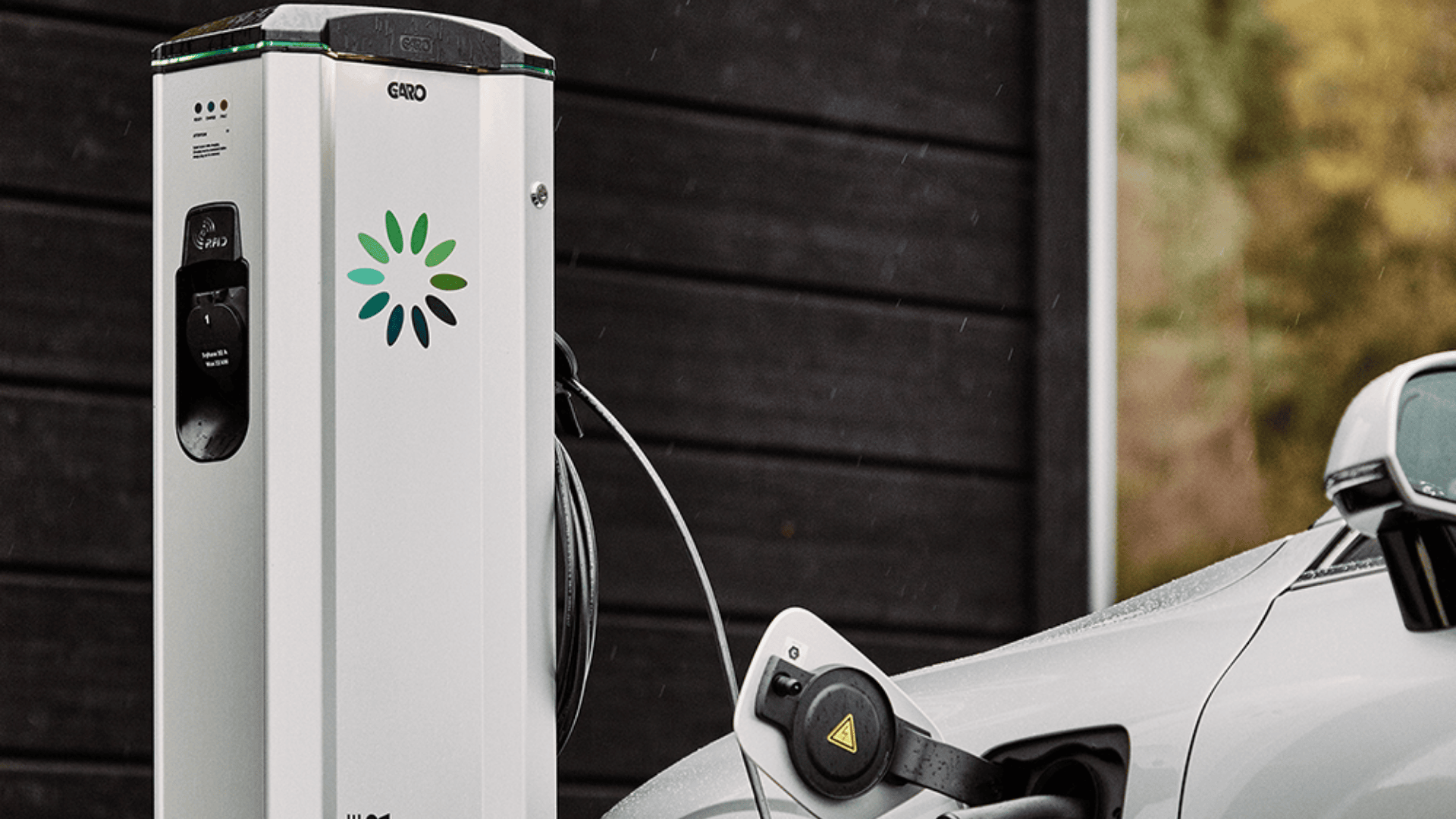 Eine Nahaufnahme eines Elektroautos, das an einer Ladestation im Freien aufgeladen wird. Das Ladekabel ist in das Auto eingesteckt, und die Station ist mit einem grünen und blauen kreisförmigen Logo versehen. Die Szene ist vor einer dunklen Holzwand zu sehen.