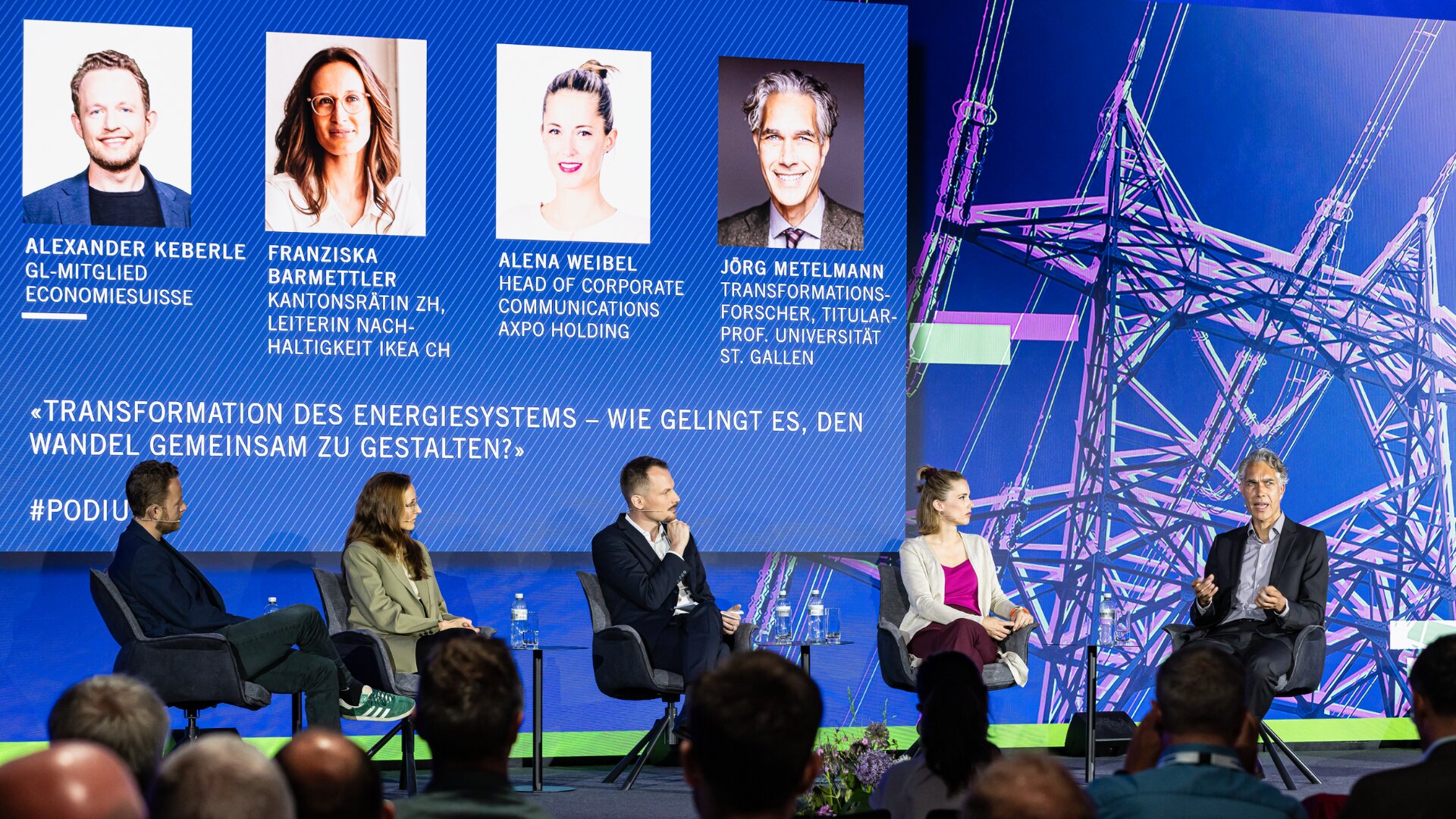 Fünf Diskussionsteilnehmer sitzen auf der Bühne vor einem Bildschirm, auf dem ihre Fotos, Namen und Titel angezeigt werden, und diskutieren auf einer Konferenz über die Energiewende. Im Hintergrund erscheint eine große Stromnetzgrafik.