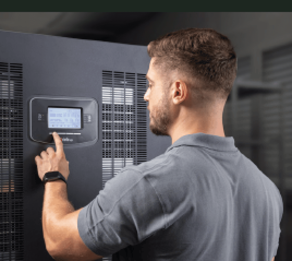 Ein Mann in einem grauen Polohemd interagiert mit einem Bedienfeld an einer großen, schwarzen Industriemaschine in einem Innenraum.