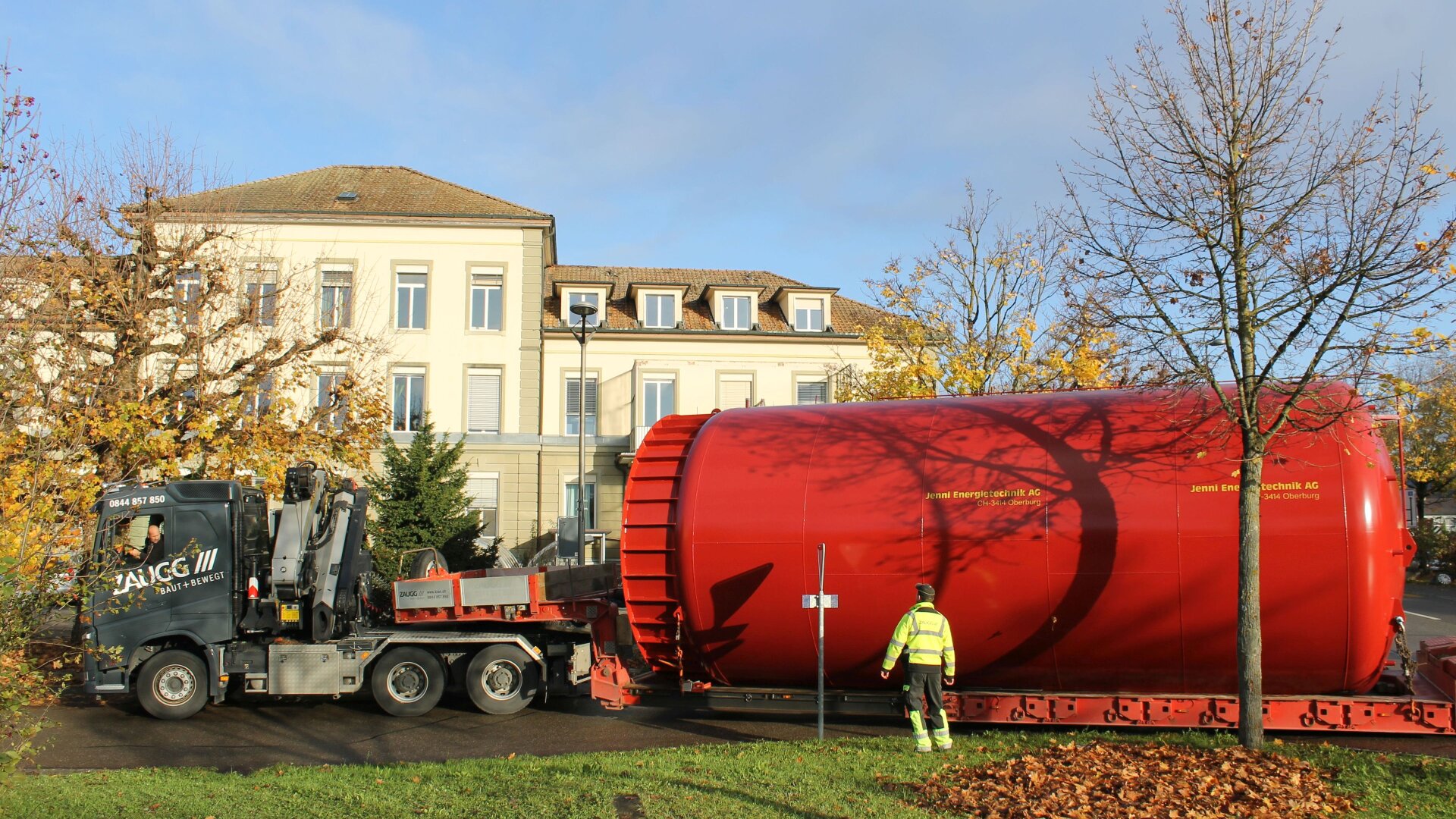 Ein großer roter zylindrischer Tank wird auf einem Tieflader transportiert. Ein Arbeiter in einem gelben Sicherheitsanzug steht in der Nähe. Im Hintergrund sind ein beigefarbenes mehrstöckiges Gebäude und Bäume mit Herbstlaub zu sehen.