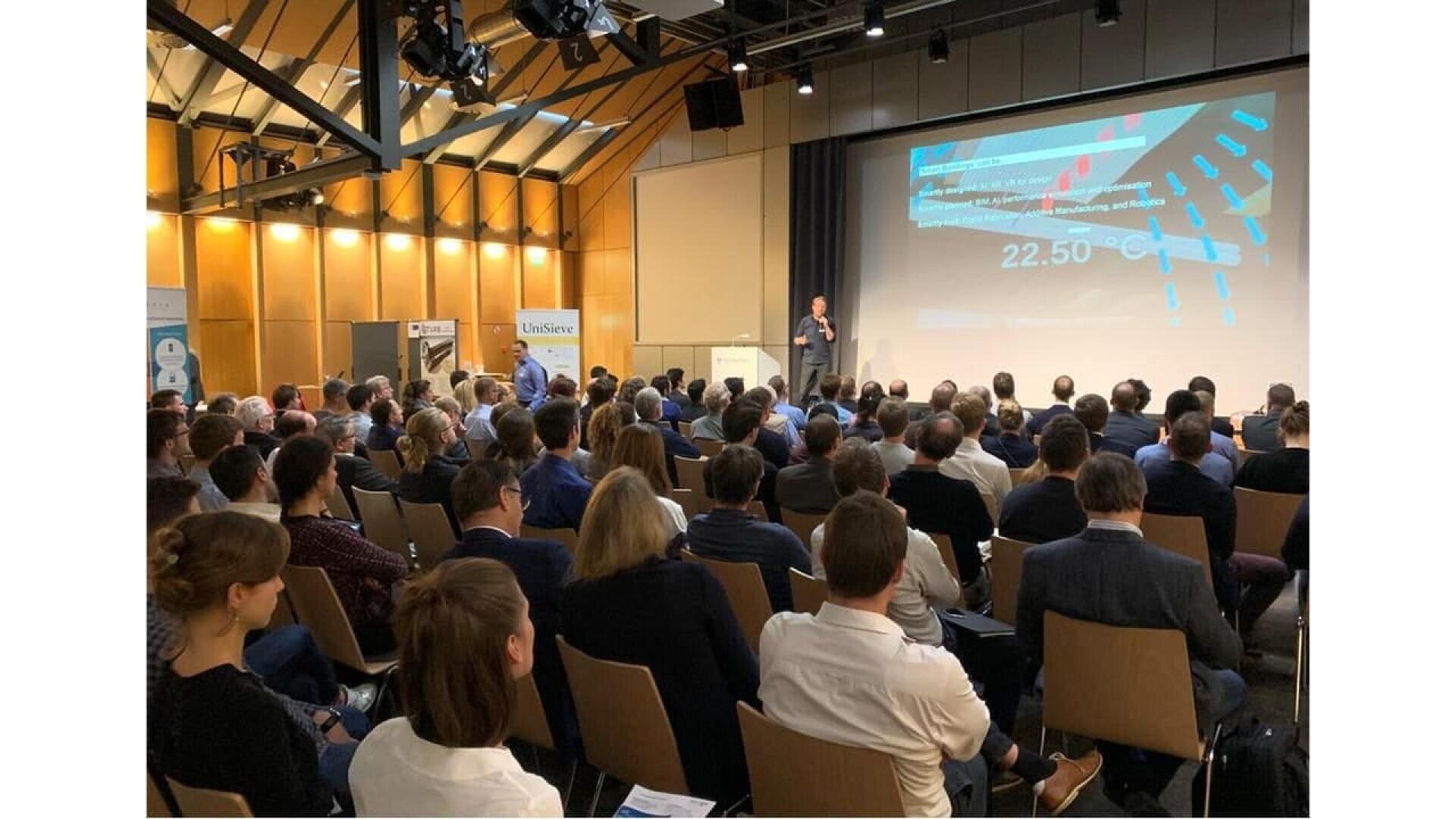 A large audience sits in rows facing a stage where a person is giving a presentation with slides projected onto a screen in a modern conference room. The room is well-lit with high ceilings and large windows.