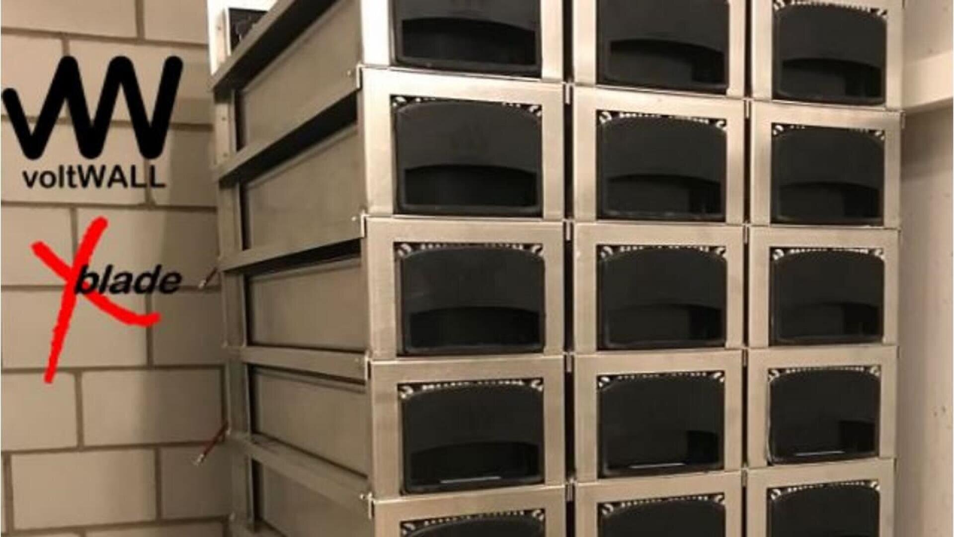 Ein Stapel metallischer Batteriemodule ist ordentlich vor einer gekachelten Wand angeordnet. Links oben ist das voltWALL-Logo zu sehen, darunter ein rot durchgestrichenes Blatt.