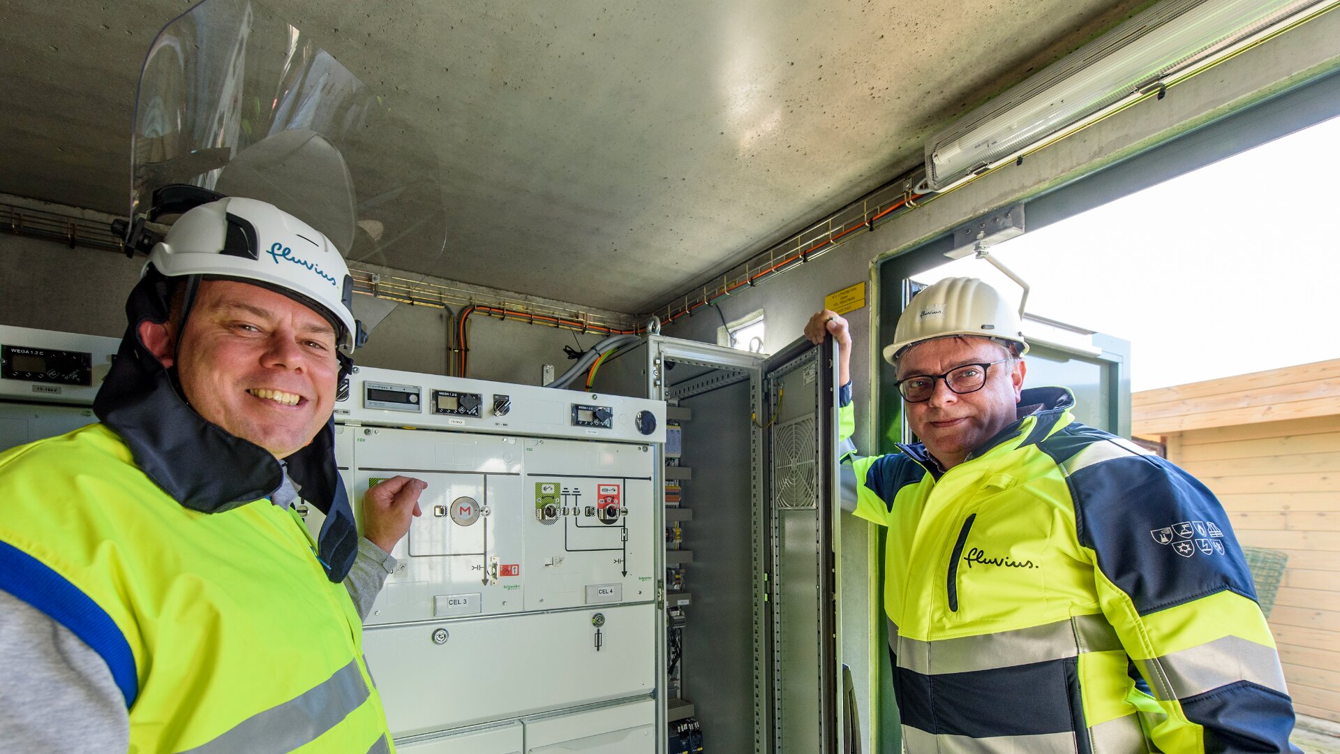 Zwei Arbeiter in Warnwesten und Schutzhelmen stehen in einem elektrischen Kontrollraum und lächeln in die Kamera. Ein Arbeiter steht neben einer offenen Schalttafel, auf der Schalter und Kabel sichtbar sind.