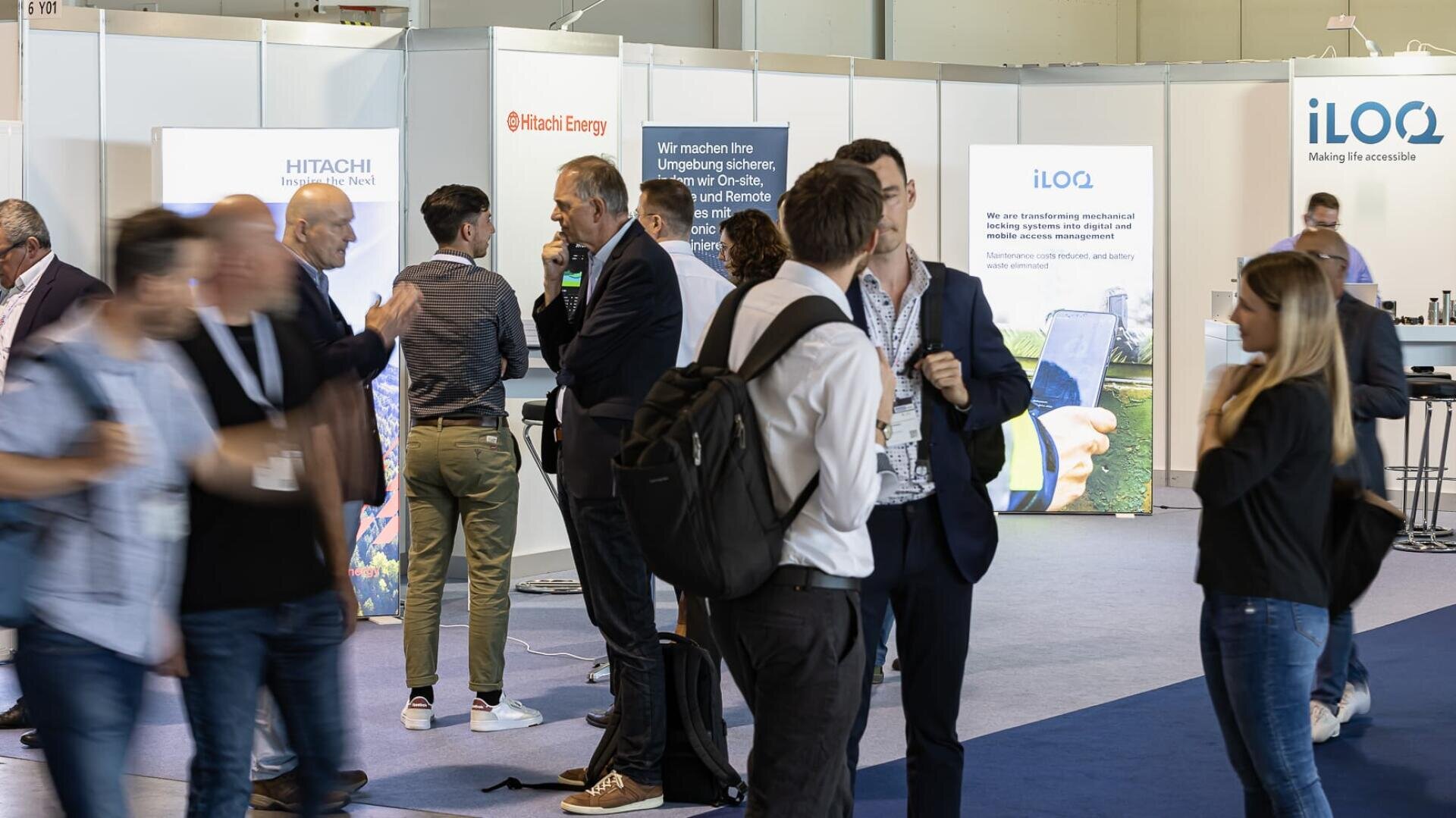 Menschen unterhalten sich auf einer Messe oder Konferenz, stehen in der Nähe von Ständen mit Firmenschildern wie Hitachi Energy und iLOQ. Einige tragen Anstecker und Rucksäcke; der Hintergrund ist hell und professionell.