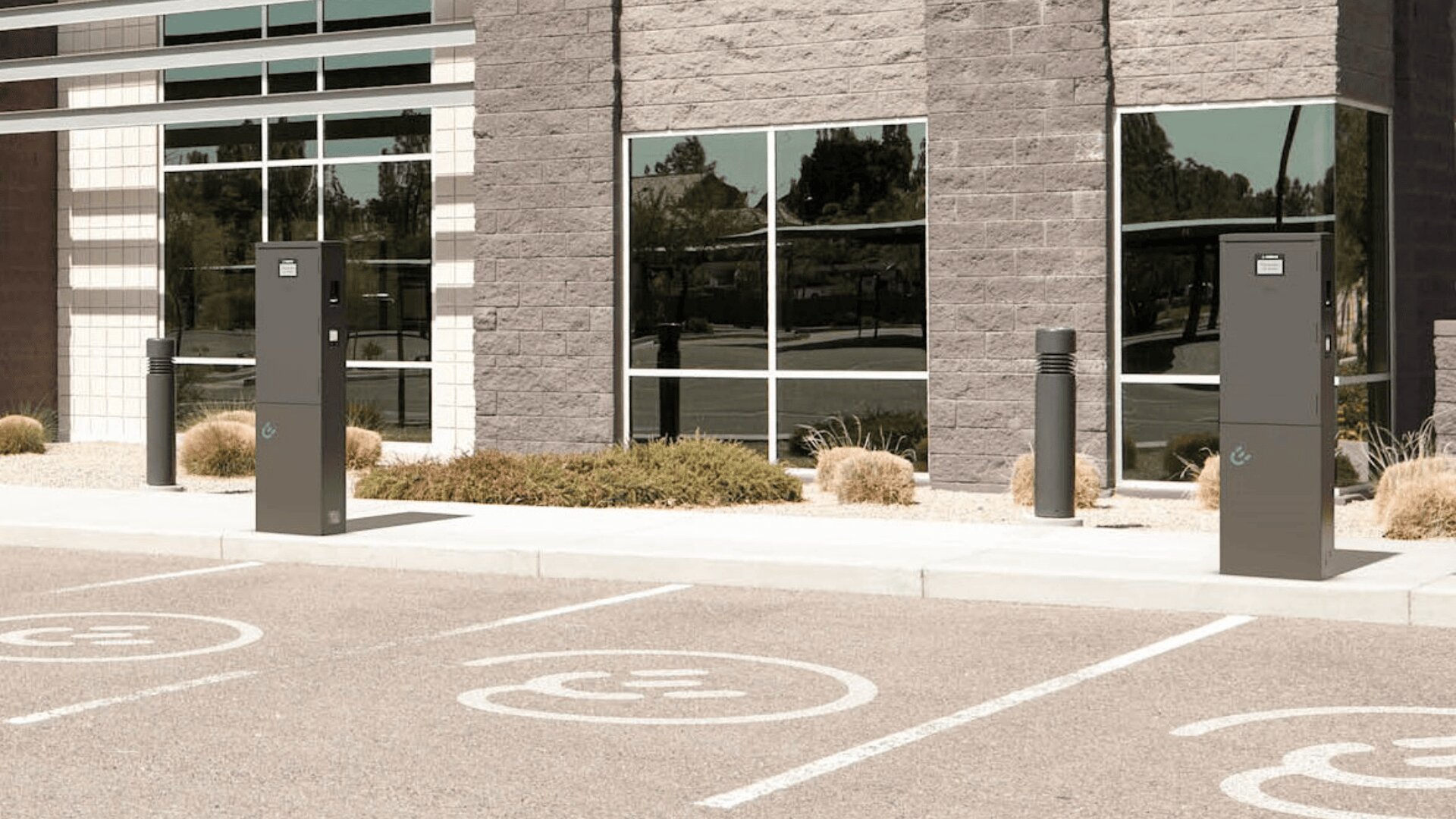 Zwei Ladestationen für Elektrofahrzeuge auf einem leeren Parkplatz vor einem modernen Gebäude mit großen Fenstern. Die Parkplätze sind mit Symbolen für Elektroautos gekennzeichnet. Sträucher und kleine Felsen säumen den Gehweg.