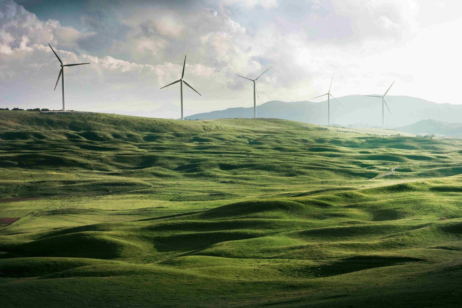 Wind turbines stand on a distant hill above rolling green fields under a partly cloudy sky, with mountains visible in the background.