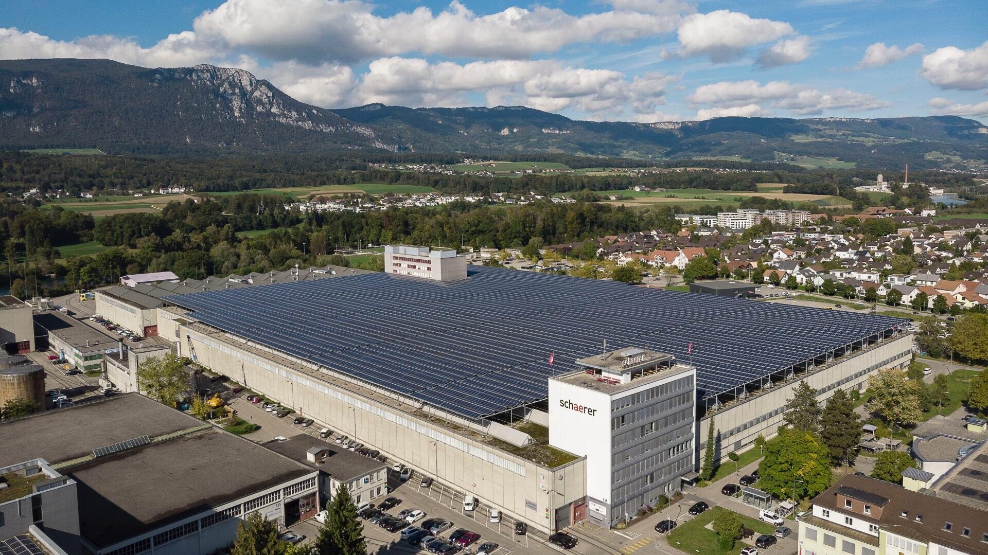Ein großes Industriegebäude mit einem mit Sonnenkollektoren bedeckten Dach, umgeben von anderen Gebäuden, Parkplätzen, Grünflächen und Bergen im Hintergrund unter einem teilweise bewölkten Himmel.