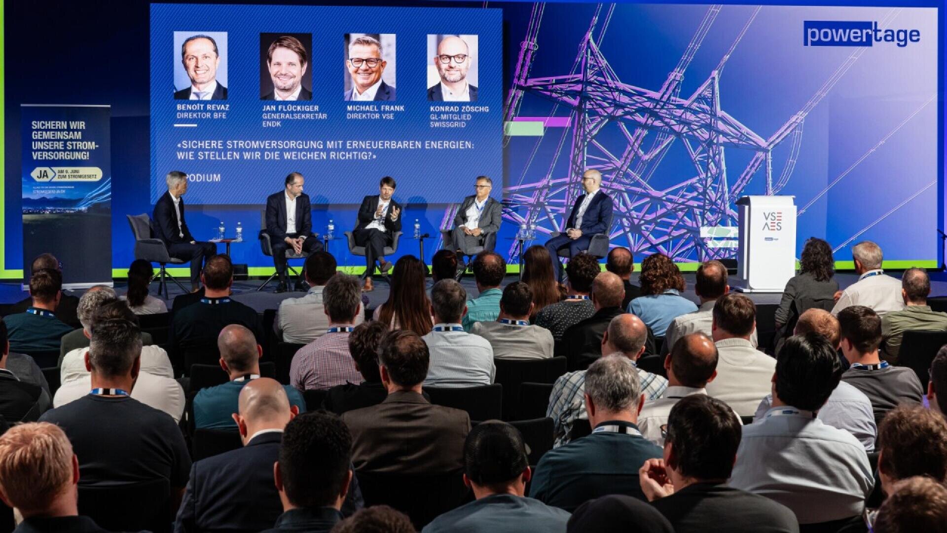 A panel of five men sits on a stage discussing energy security at a conference, with a large audience watching. A screen behind them displays their names, photos, and the event title in German, alongside a power grid image.