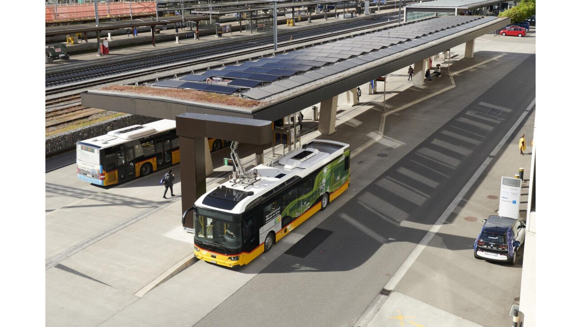 An electric bus charges at a station under a solar panel canopy near train tracks. Another bus and a car are parked nearby. The scene is bright and modern, with clear lines and a clean, organized layout.