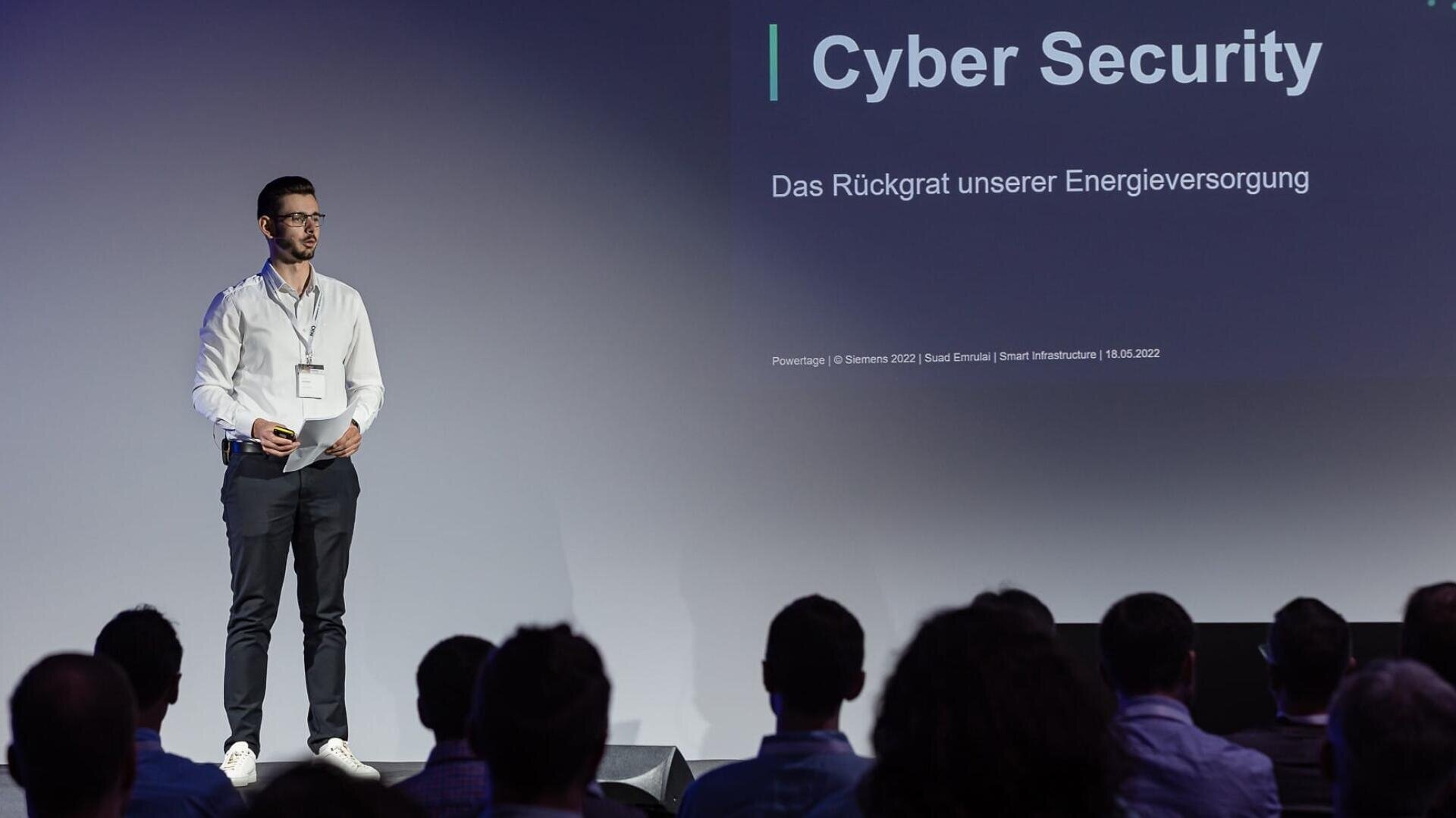 Ein Mann steht auf der Bühne und hält einen Vortrag über Cybersicherheit. Auf der Folie hinter ihm steht "Cybersicherheit: Das Rückgrat unserer Energieversorgung". Ein Publikum schaut aufmerksam zu.
