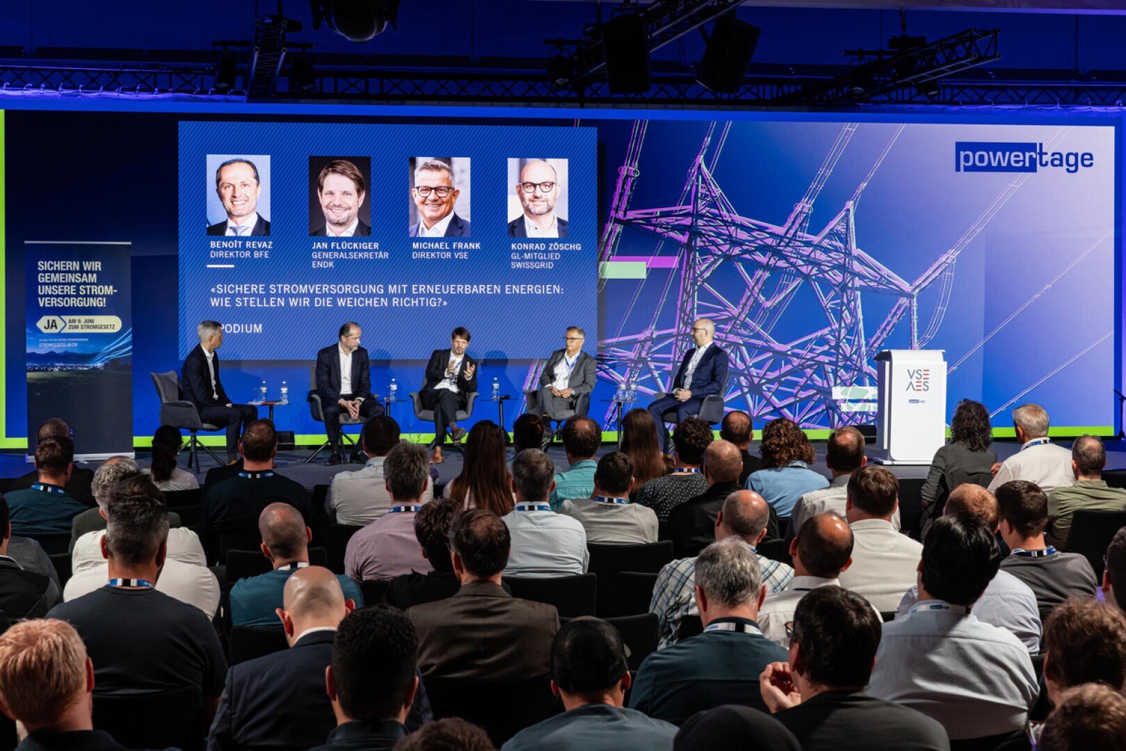 Panel discussion at a conference where five speakers sit on stage in front of a large audience. Behind them is a screen on which their names, photos and the topic of sustainable energy can be seen.