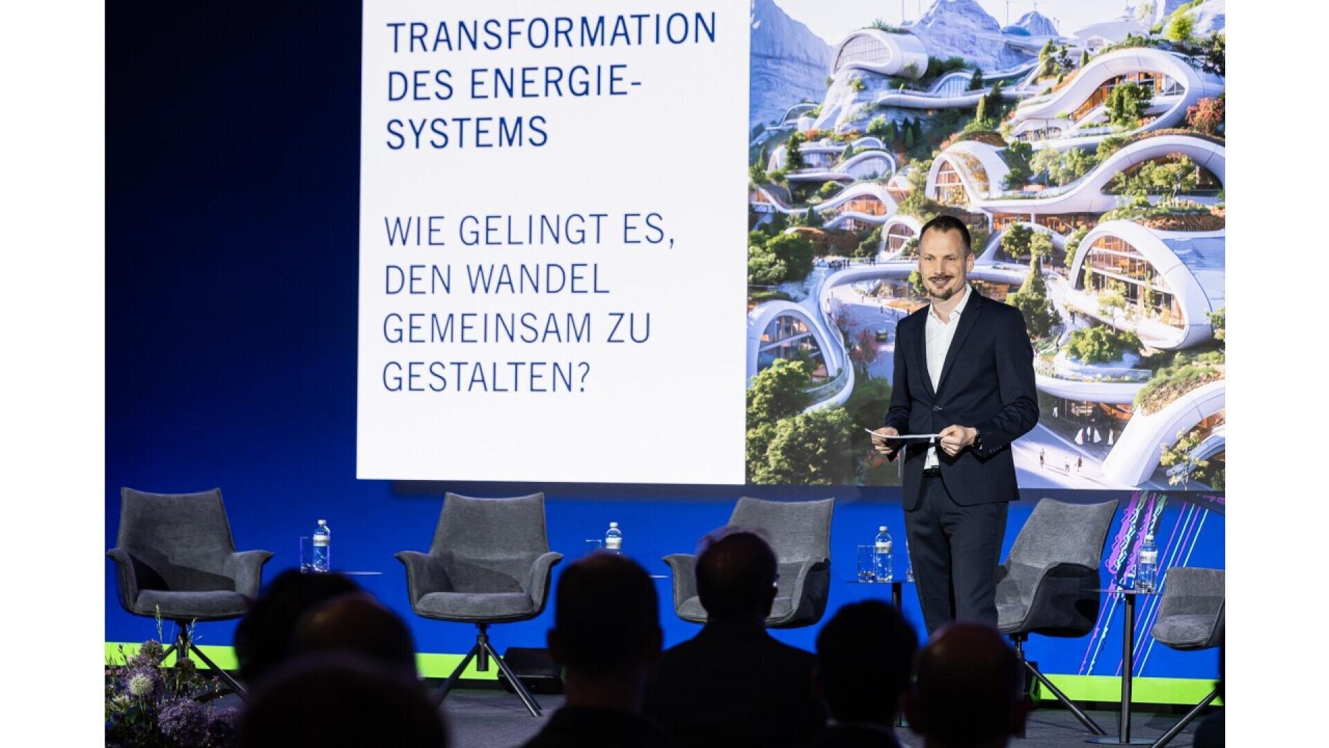 A man in a suit stands on stage with a microphone and tablet, speaking in front of a screen displaying German text about energy transformation; several empty chairs and water bottles are visible.