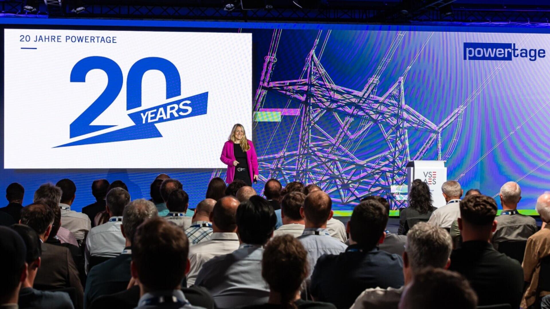 A woman speaks on stage to an audience at an event celebrating 20 Years Powertage. A large screen displays the anniversary logo, and a blue and purple graphic appears in the background.