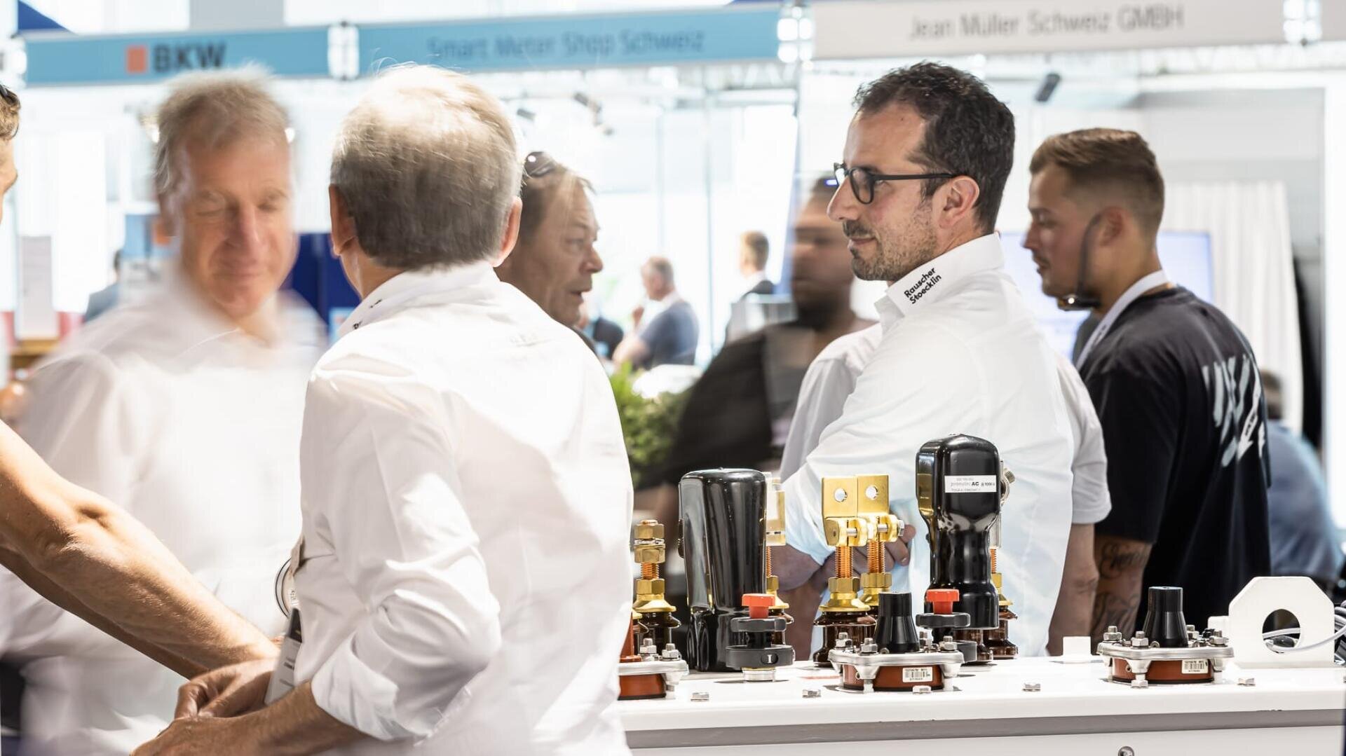 Eine Gruppe von Männern in weißen Hemden unterhält sich auf einer Technologie-Messe. Im Vordergrund sind verschiedene elektrische Komponenten auf einem Tisch ausgestellt. Im unscharfen Hintergrund sind weitere Personen und Firmenschilder zu sehen.