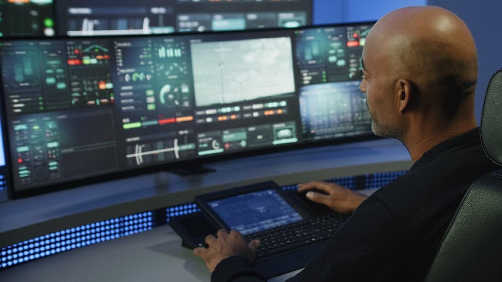 A person sits at a control desk with multiple screens displaying various technical data and images, monitoring and operating complex systems in a high-tech environment.