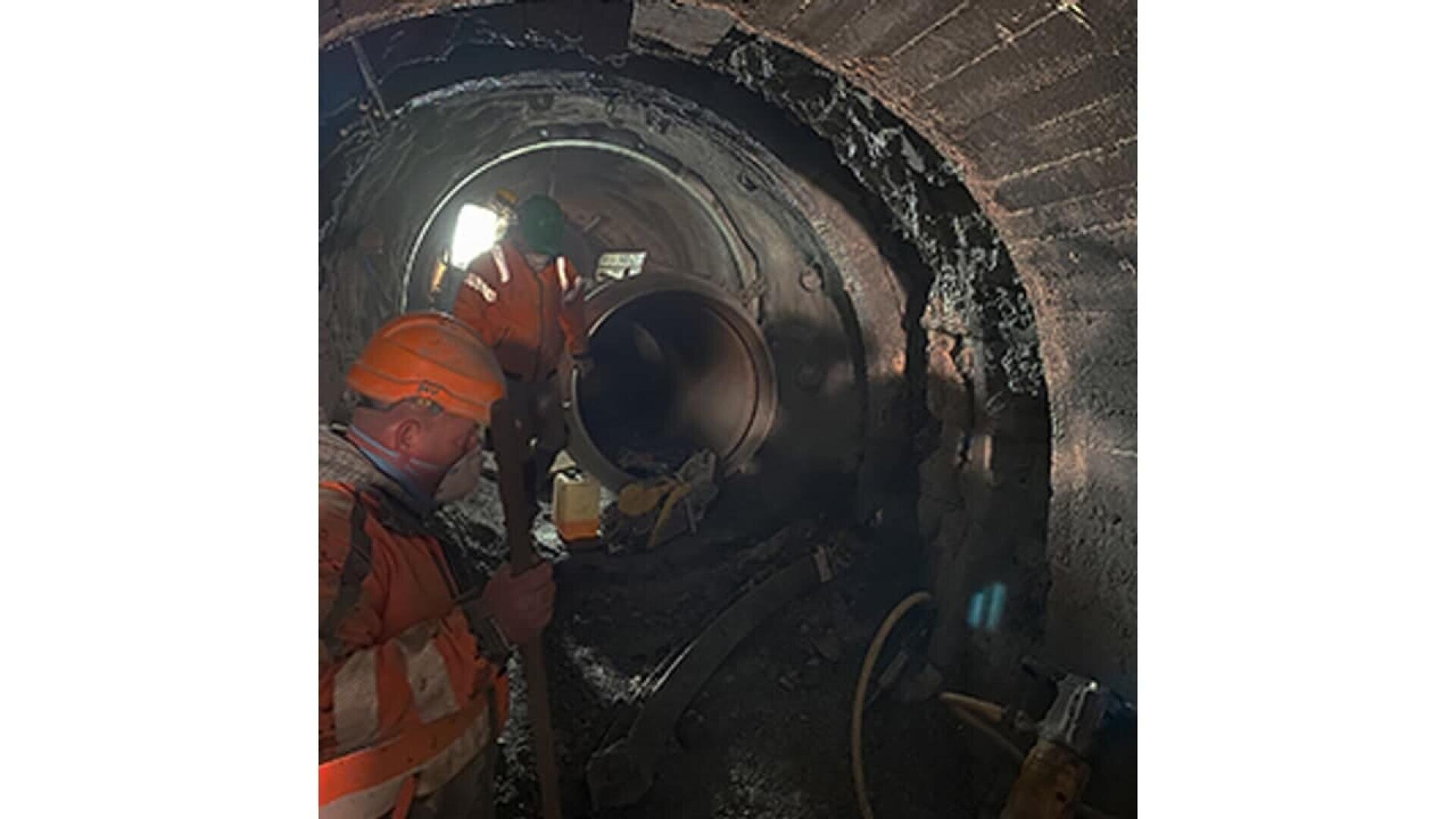 Two construction workers in orange safety gear and helmets work inside a large, dimly lit, circular tunnel. One holds a tool while the other is near a metal pipe section. The tunnel walls appear rough and darkened.