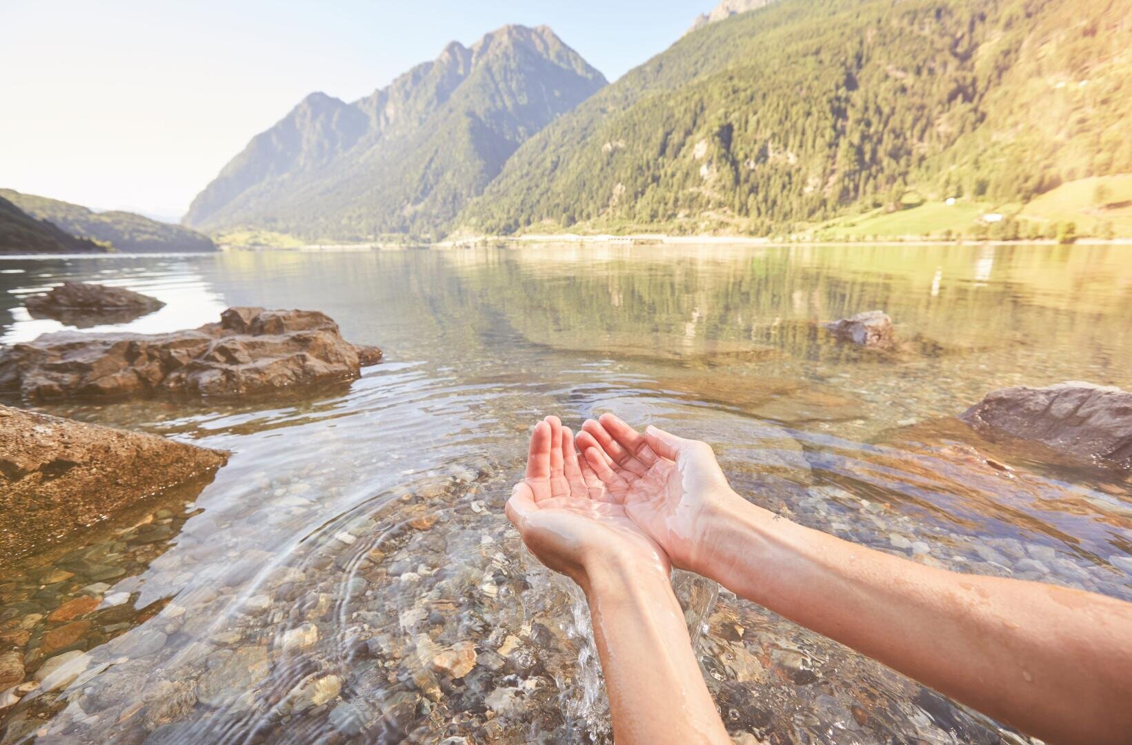 Eine Person schöpft mit ihren Händen klares Wasser aus einem ruhigen Bergsee, der von Felsen und bewaldeten Hügeln umgeben ist, mit fernen Bergen unter einem hellen Himmel.