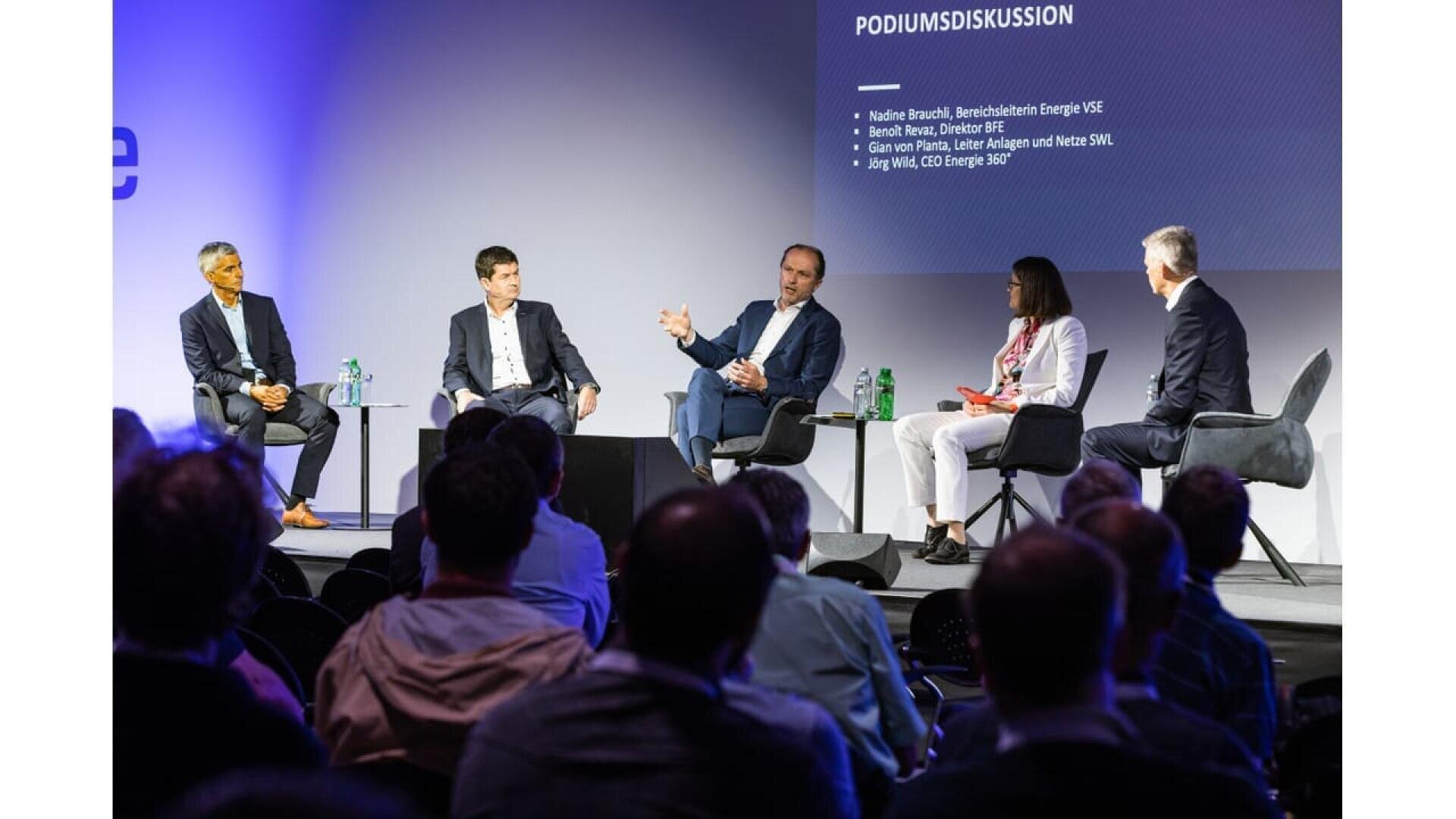 Five people in business attire sit on a stage in a panel discussion, speaking to an audience. A screen behind them displays information in German, with names and job titles of the panelists.