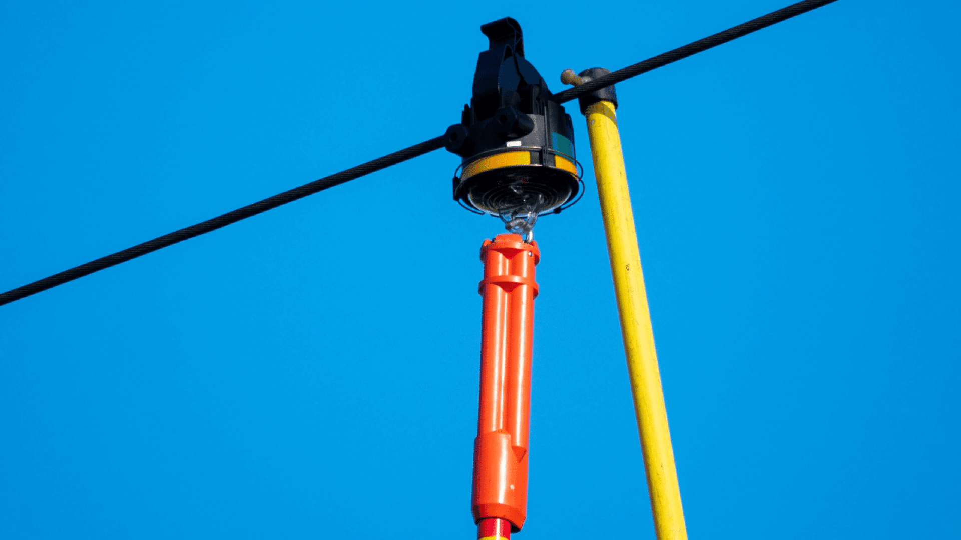 Ein leuchtend orangefarbenes und gelbes Leitungswerkzeug wird vor dem Hintergrund eines strahlend blauen Himmels an einer Freileitung befestigt und demonstriert die Ausrüstung für die sichere Arbeit mit elektrischen Leitungen.