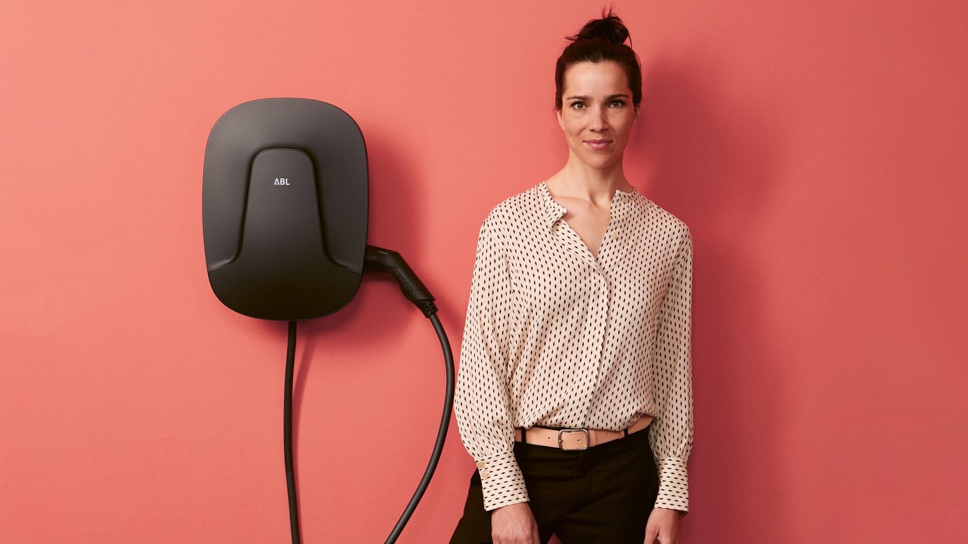 Eine Frau mit dunklem Haar in einem Dutt steht an einer korallenfarbenen Wand neben einer schwarzen Ladestation für Elektrofahrzeuge, die an der Wand angebracht ist. Sie trägt eine helle Bluse mit einem Muster und eine dunkle Hose.