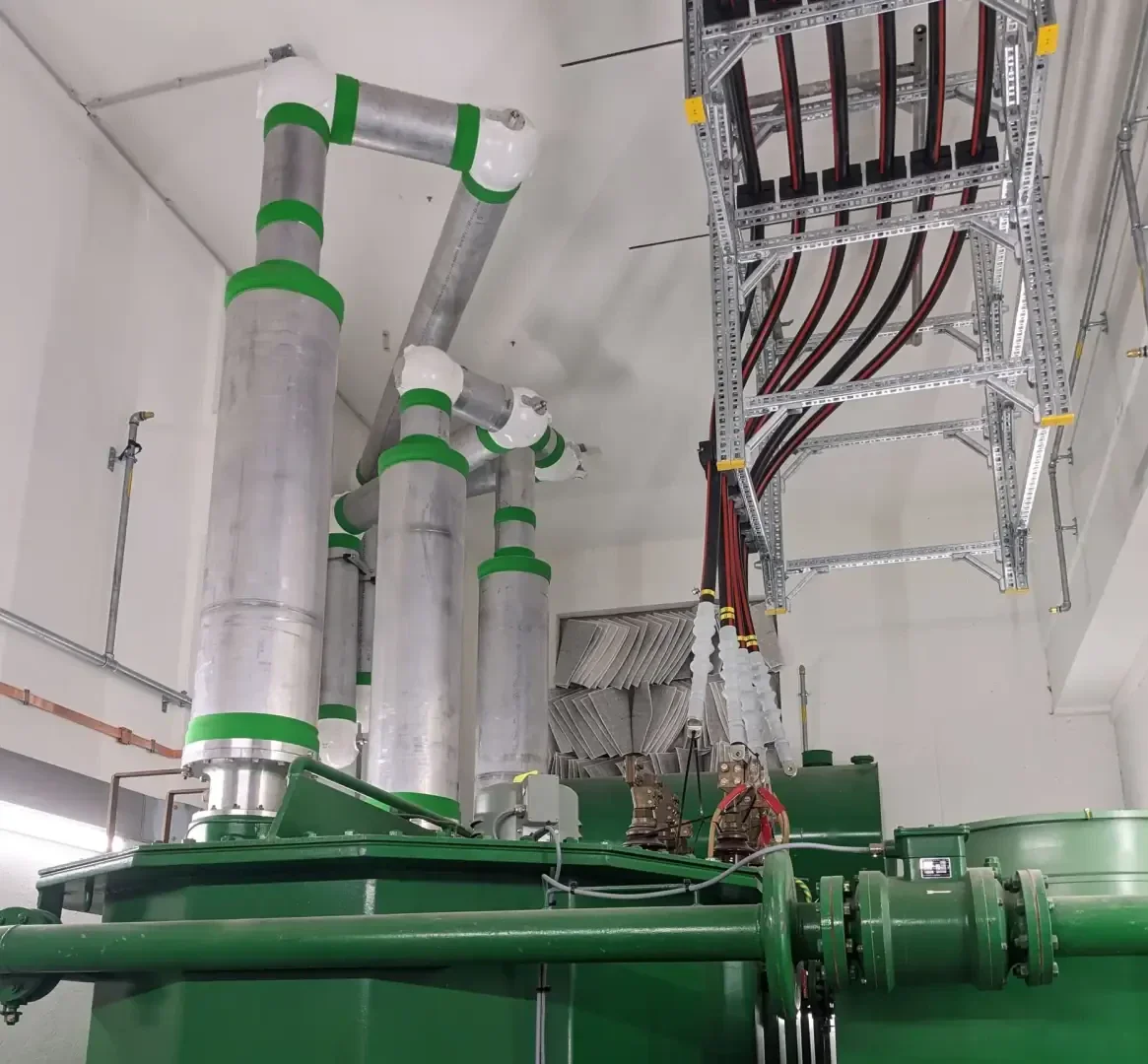 Large silver pipes with green bands are connected to green industrial machines in a clean, white room. Above the head, a metal cable tray carries several red and black cables.
