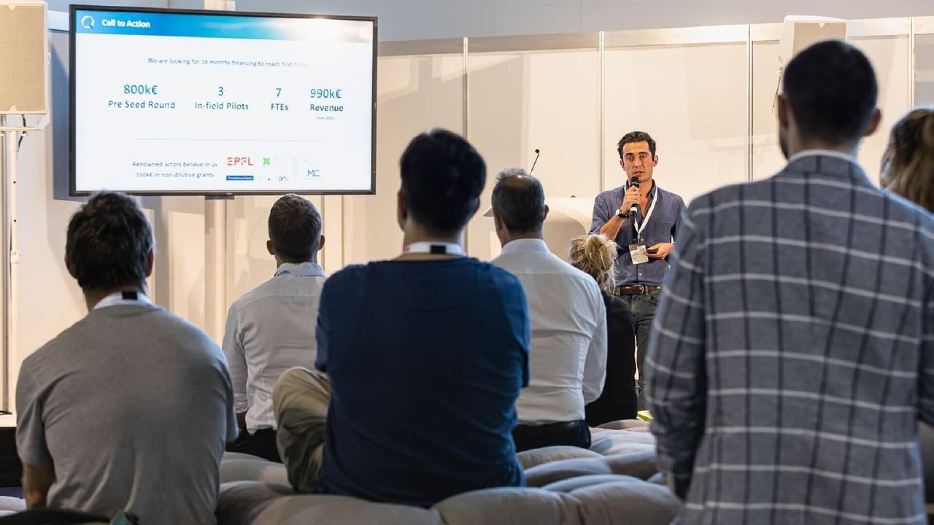A man stands speaking into a microphone and presenting slides to an audience seated indoors. The slide behind him displays funding, pilot, and revenue figures, while listeners focus on the presentation.