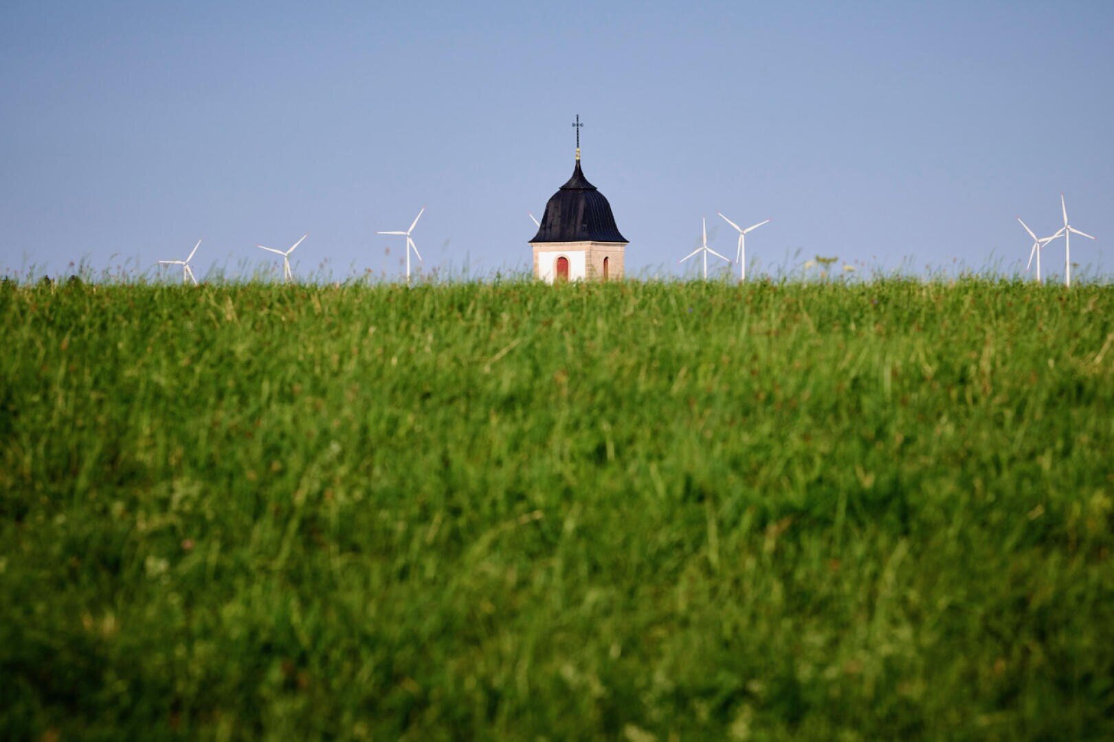 A church tower with a cross rises above a lush green field, with several wind turbines visible in the background against a clear blue sky.