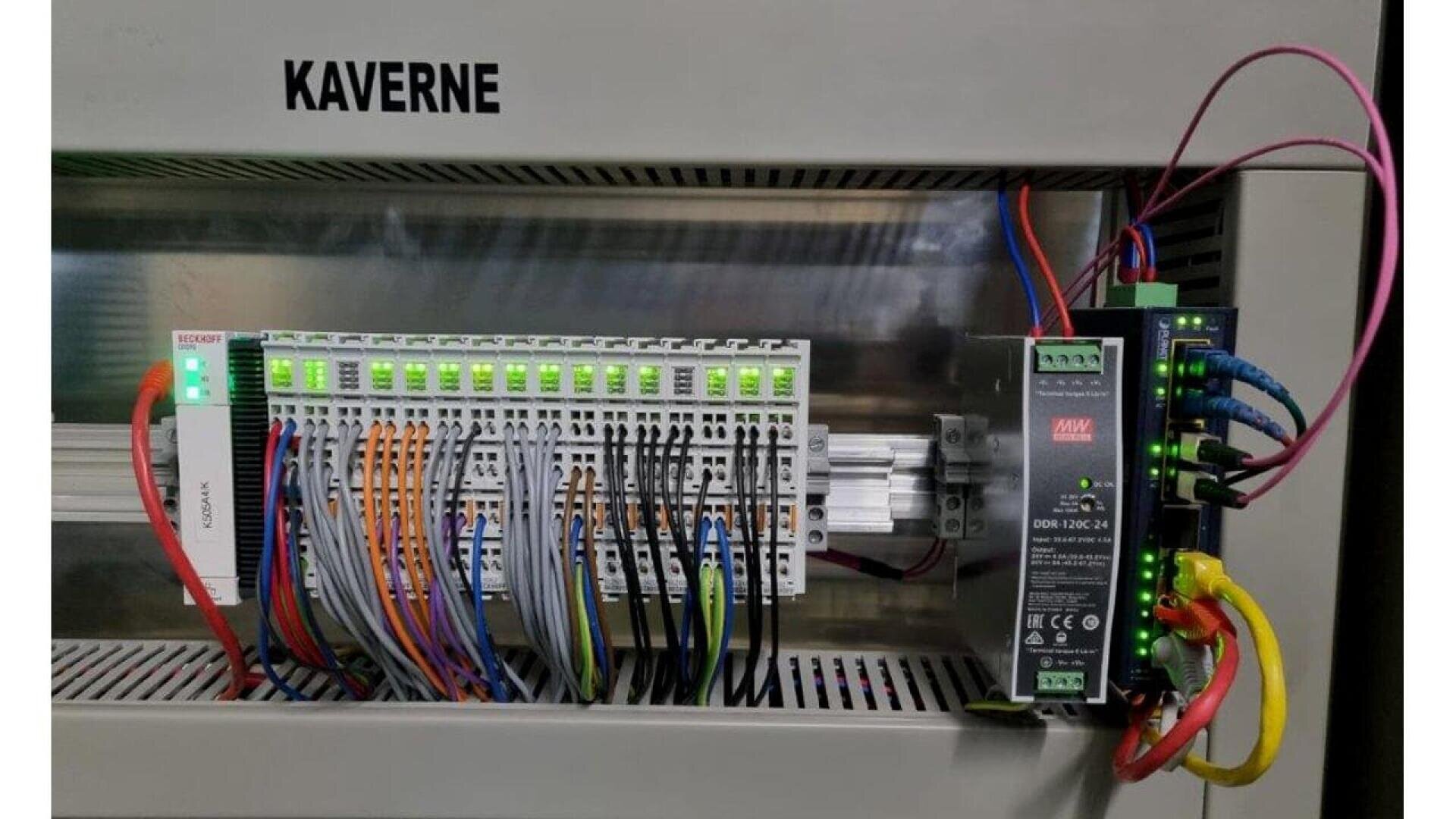 A control panel with labeled KAVERNE, showing neatly organized terminal blocks, numerous colored wires, illuminated green indicator lights, and electronic modules inside an industrial enclosure.