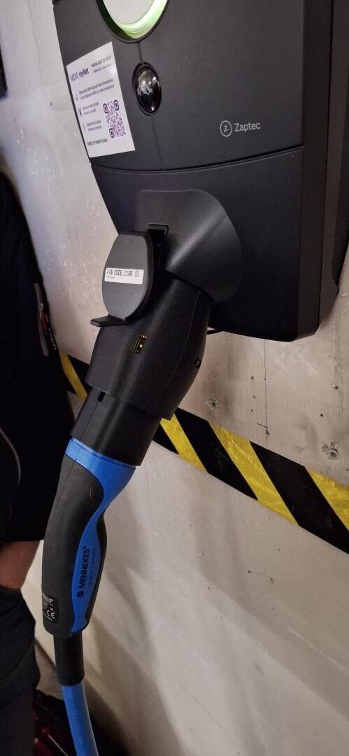 A close-up of a blue and black electric vehicle charging plug connected to a Zaptec charging station mounted on a white wall with black and yellow caution tape.