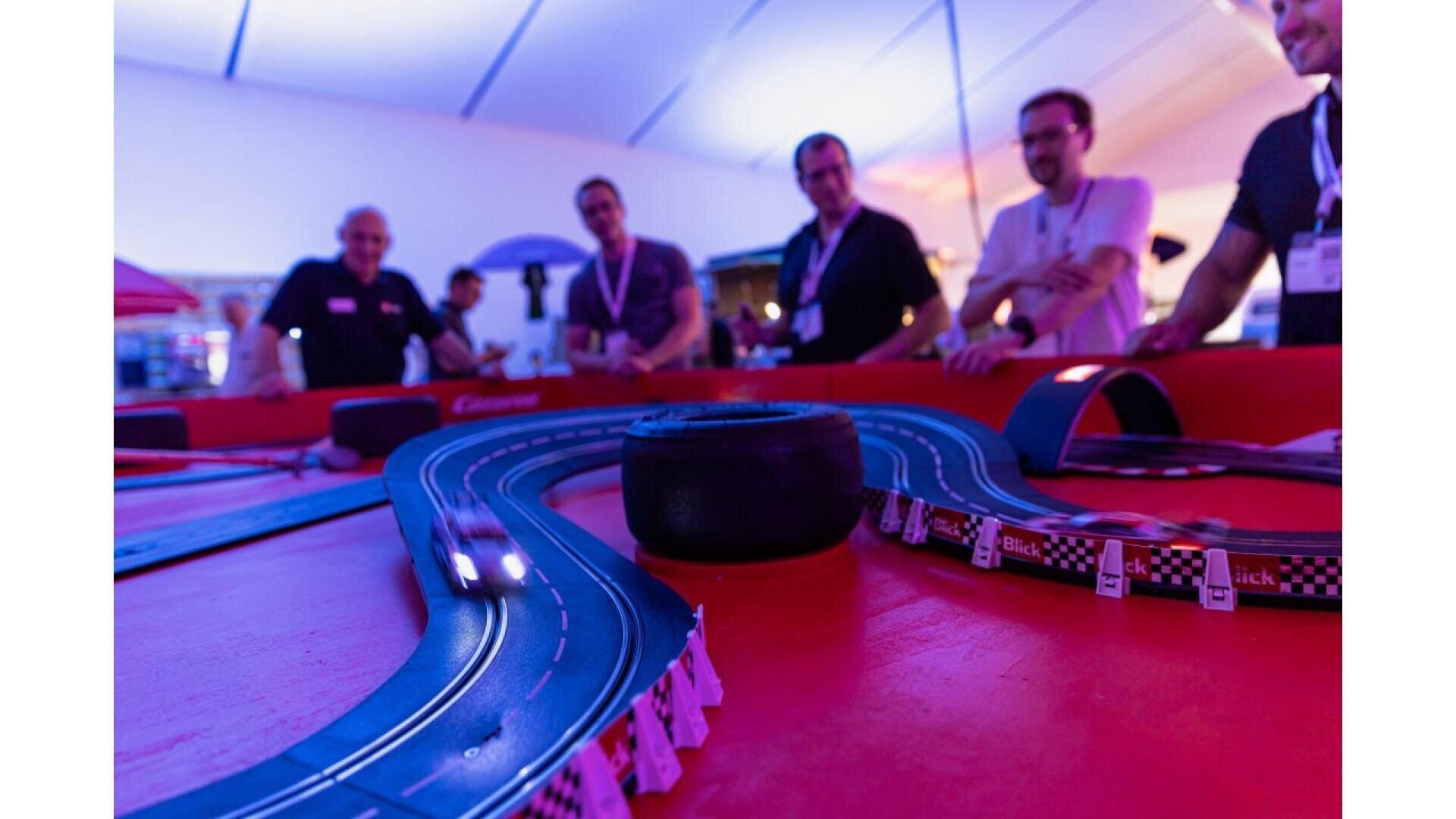 A group of casually dressed people stand around a small race car track, watching a toy car speed along the winding lanes under purple and blue lighting. The atmosphere is lively and playful.