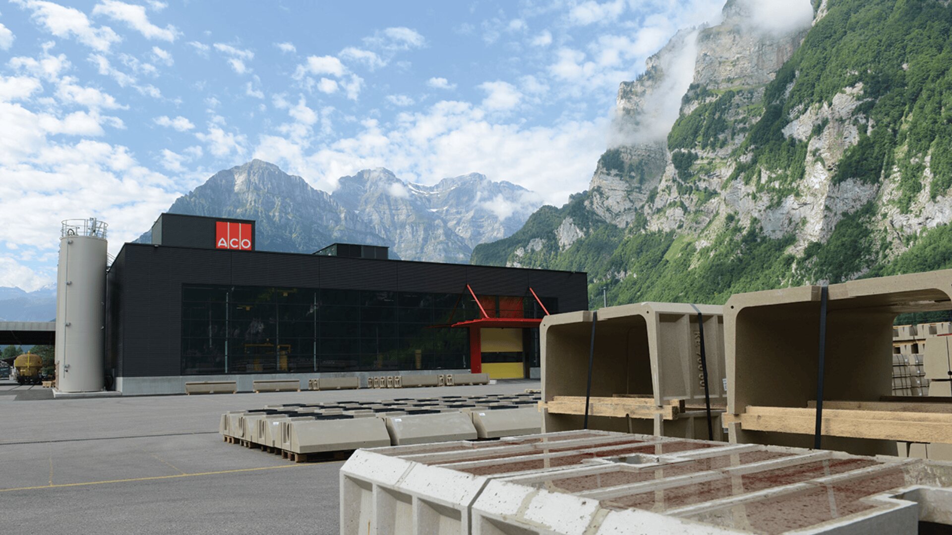 A modern industrial building with an ACO sign is surrounded by concrete products in an outdoor yard, set against a backdrop of tall, green mountains and partly cloudy blue sky.