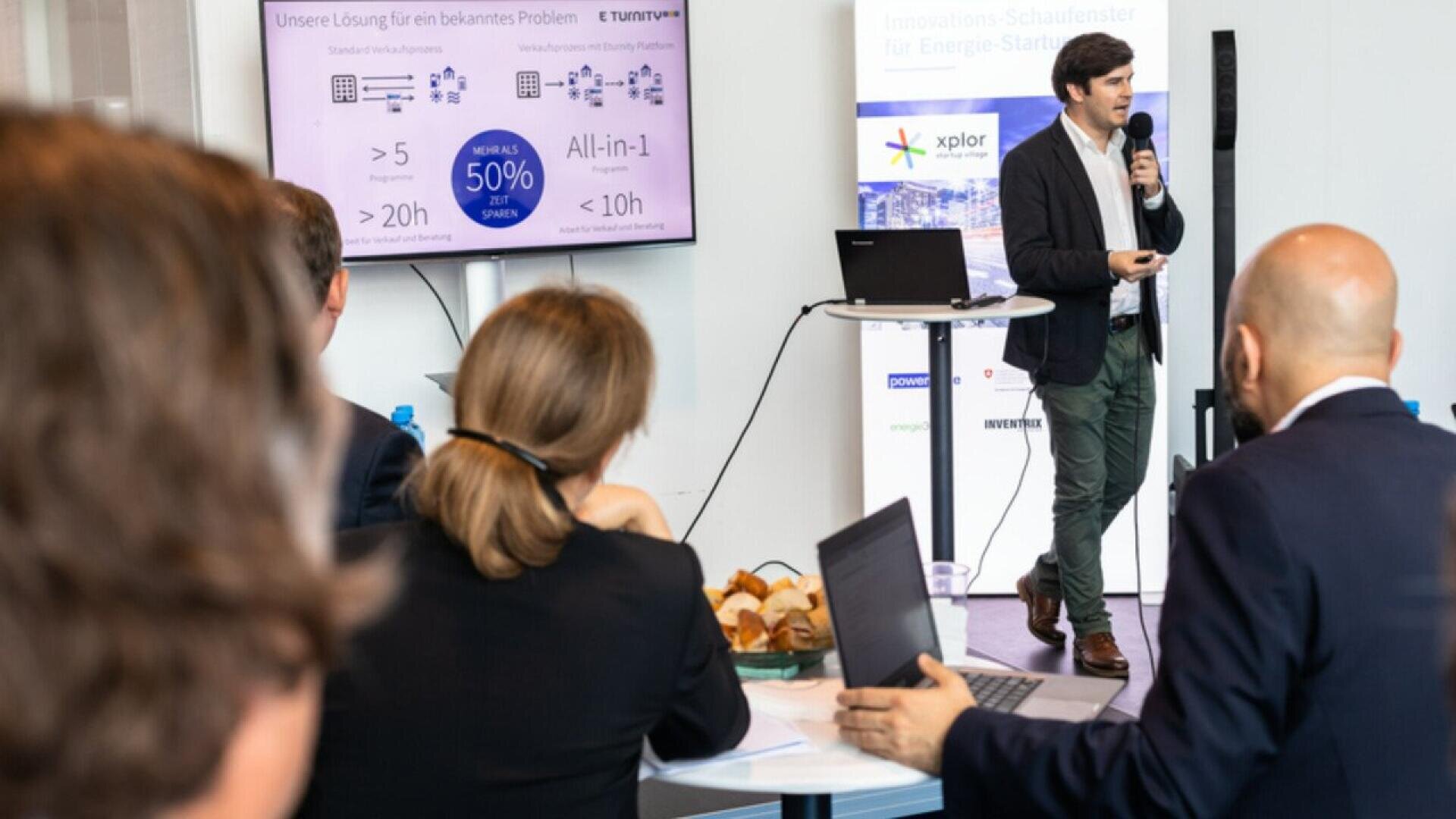 A man in business attire gives a presentation using a microphone, while a group of people sit at tables with laptops and pastries, watching a screen displaying charts and text.