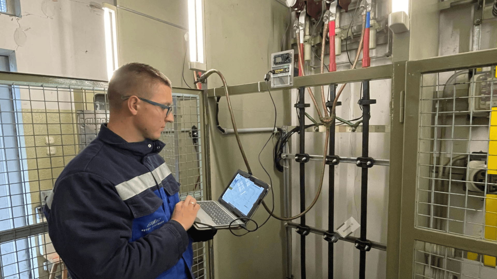 Ein Mann mit Schutzbrille und blauer Arbeitsjacke arbeitet mit einem Laptop, während er in einem eingezäunten Bereich mit Kabeln und Messgeräten elektrische Geräte überprüft.