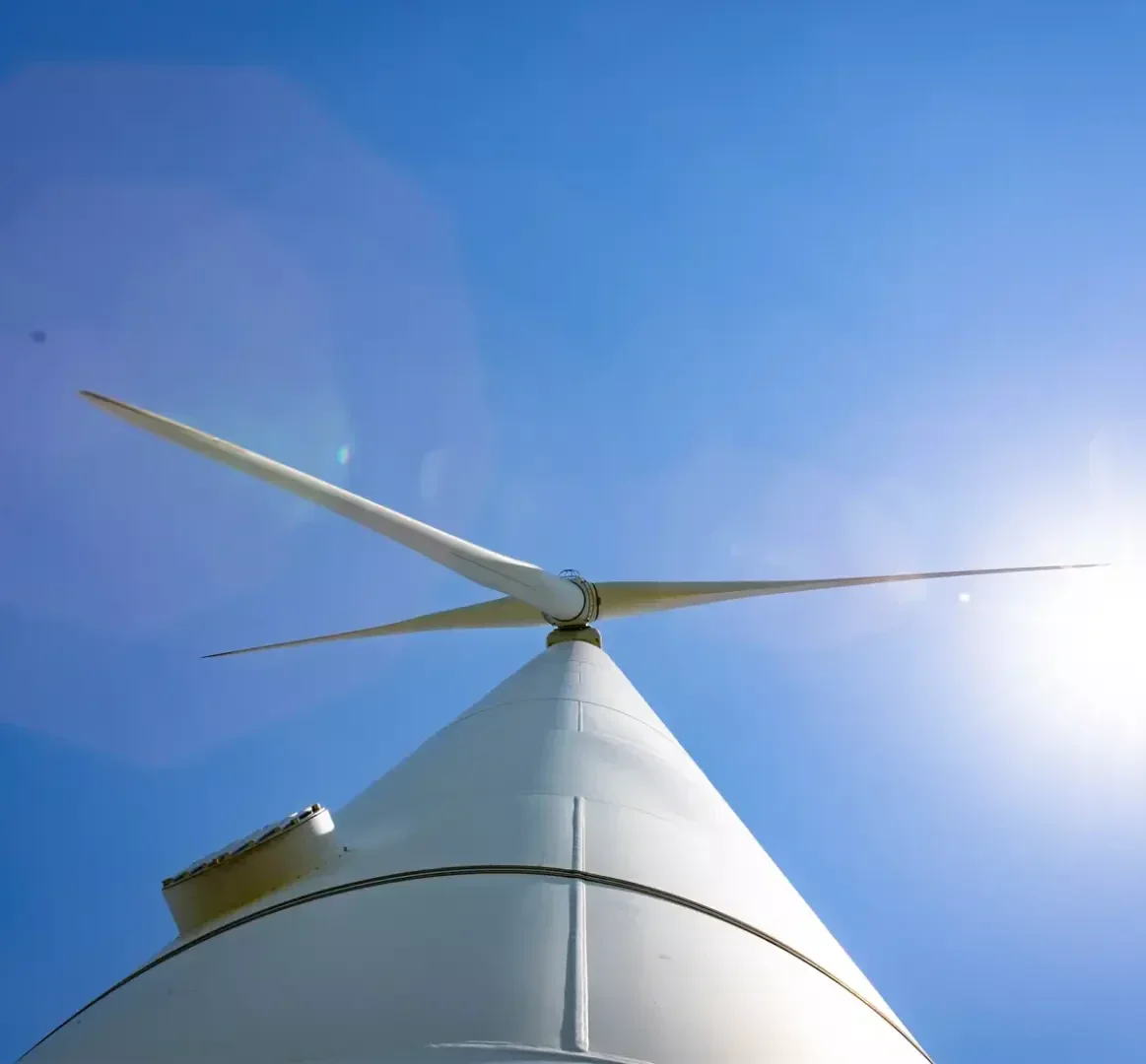 Looking up at a white wind turbine against a clear blue sky with the sun shining brightly in the background.