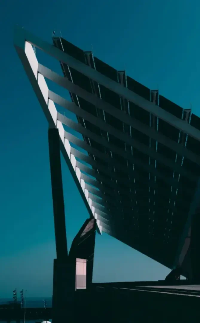 Ein großes, modernes Solarmodulfeld mit abgewinkelten, rechteckigen Paneelen steht auf stabilen Stützen vor einem klaren, blauen Himmel und wirft scharfe, geometrische Schatten nach unten.