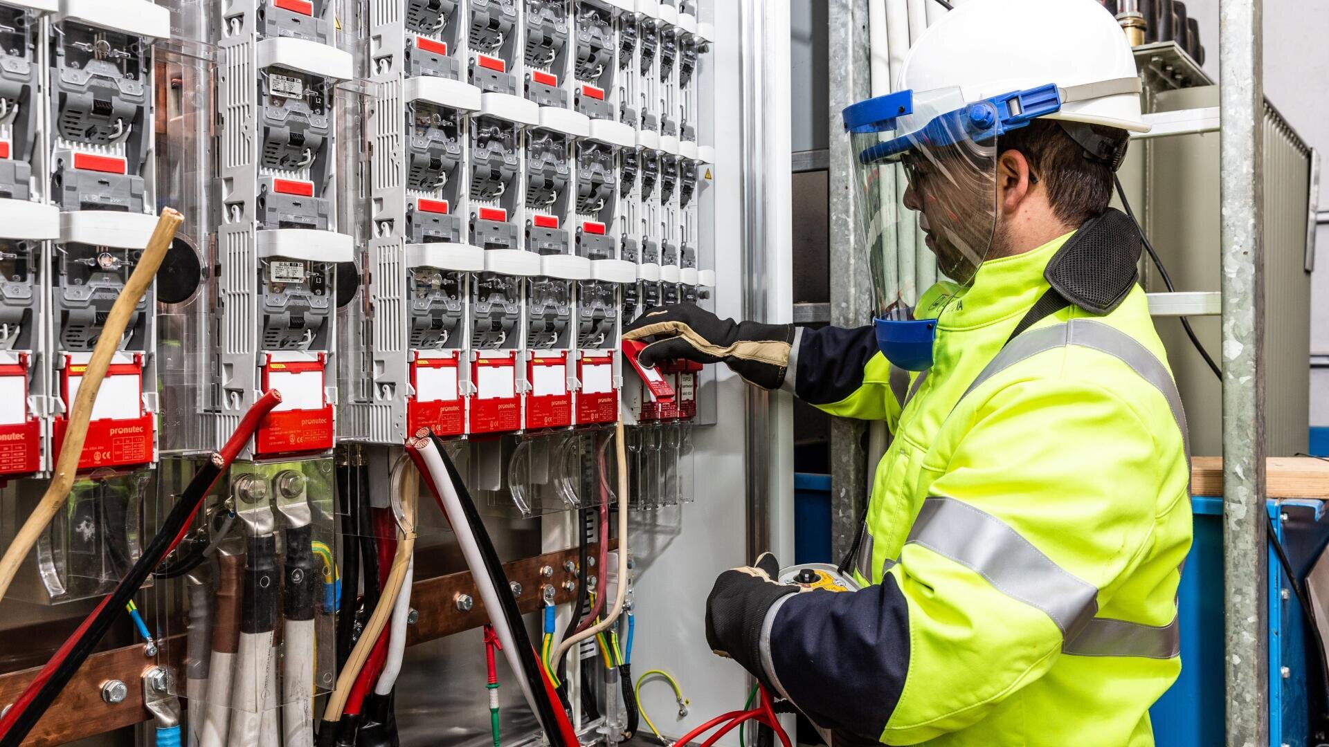 An electrician wearing a hard hat, face shield and high visibility vest is working on an electrical panel with multiple circuit breakers and red signs in an industrial environment.