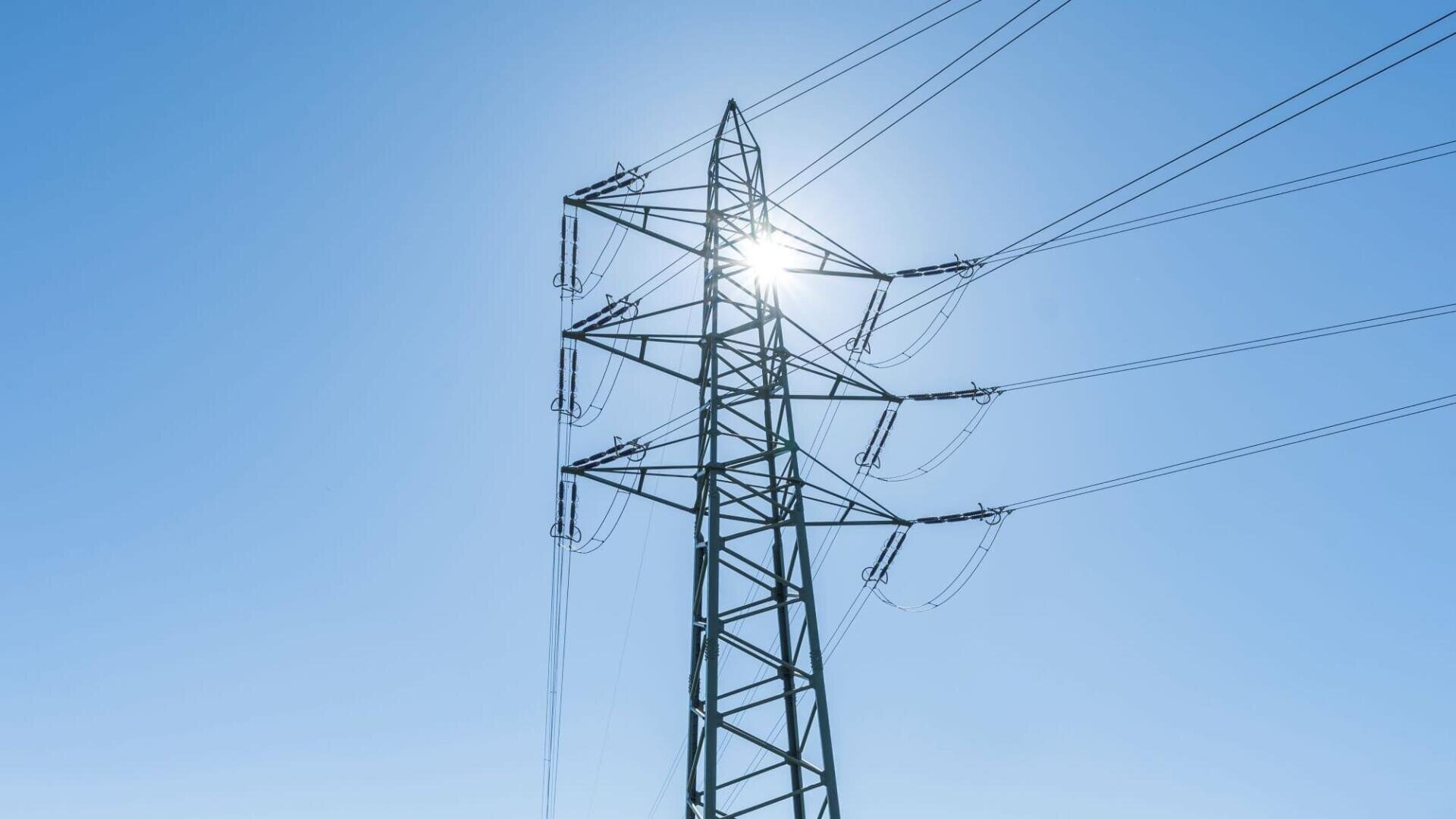 Ein hoher Hochspannungsmast mit Stromleitungen ragt in den strahlend blauen Himmel, und die Sonne scheint hell hinter der Metallstruktur des Turms.