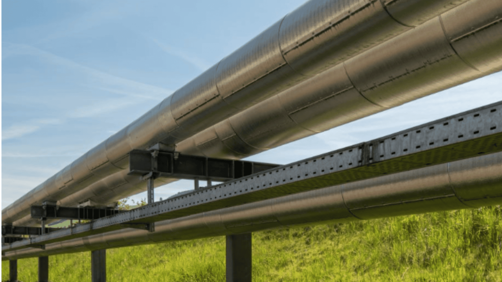 Zwei große Metallpipelines verlaufen parallel über dem Boden, gestützt von Metallkonstruktionen, mit grünem Gras darunter und einem blauen Himmel mit leichten Wolken im Hintergrund.