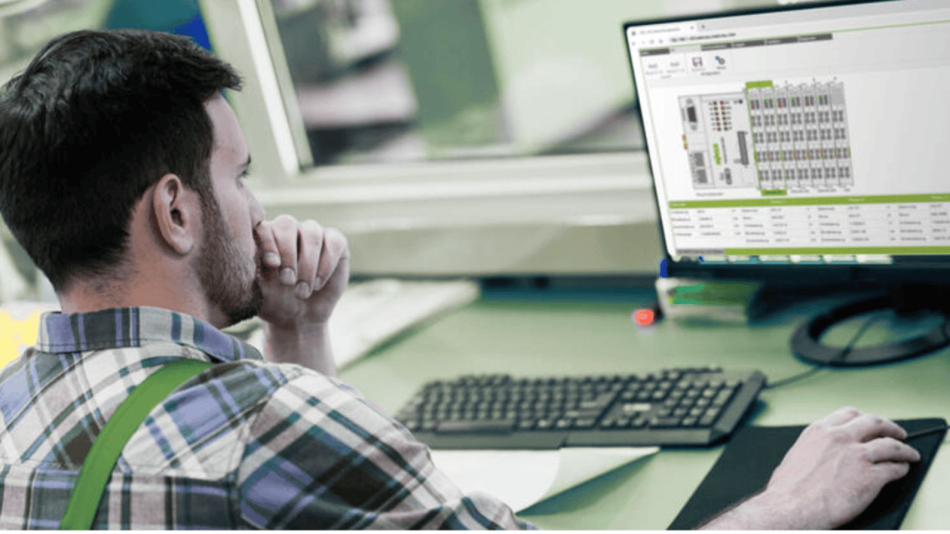 A man wearing a plaid shirt sits at a desk, concentrating on a computer screen displaying technical diagrams and data. A keyboard, mouse, and papers are on the green desk in front of him.