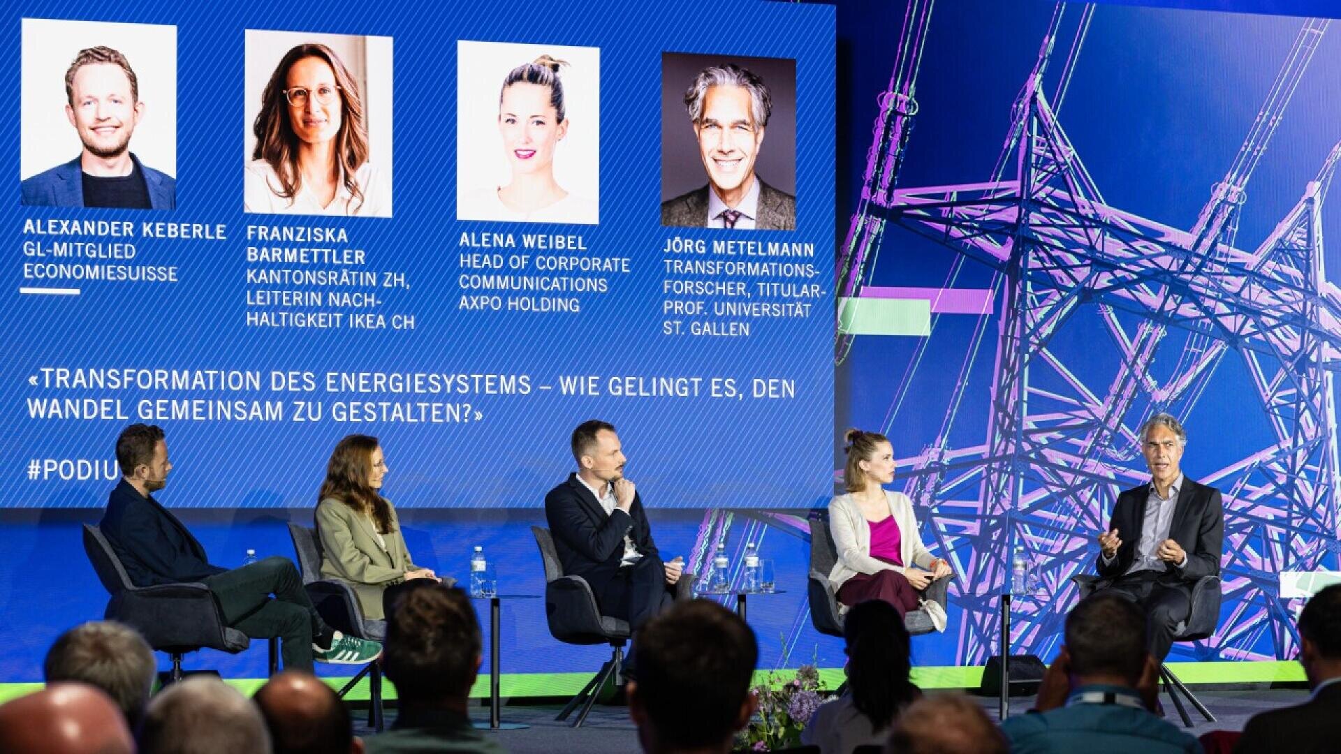 Five people sit on a panel in front of a blue screen displaying their names, titles, and photos. They are discussing the transformation of energy systems. The audience is visible in the foreground.