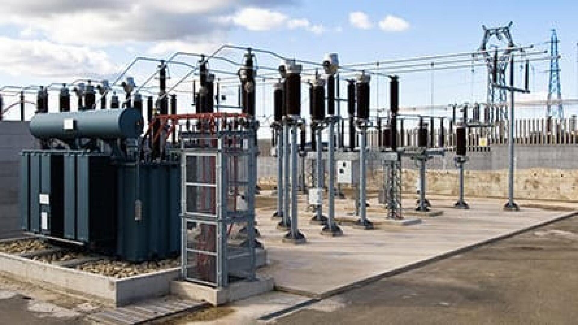 Elektrisches Umspannwerk im Freien mit Transformatoren, Leistungsschaltern und Stromleitungen auf einer Betonplattform, umgeben von einem Zaun. Der Himmel ist teilweise bewölkt, und im Hintergrund sind Industrieanlagen zu sehen.