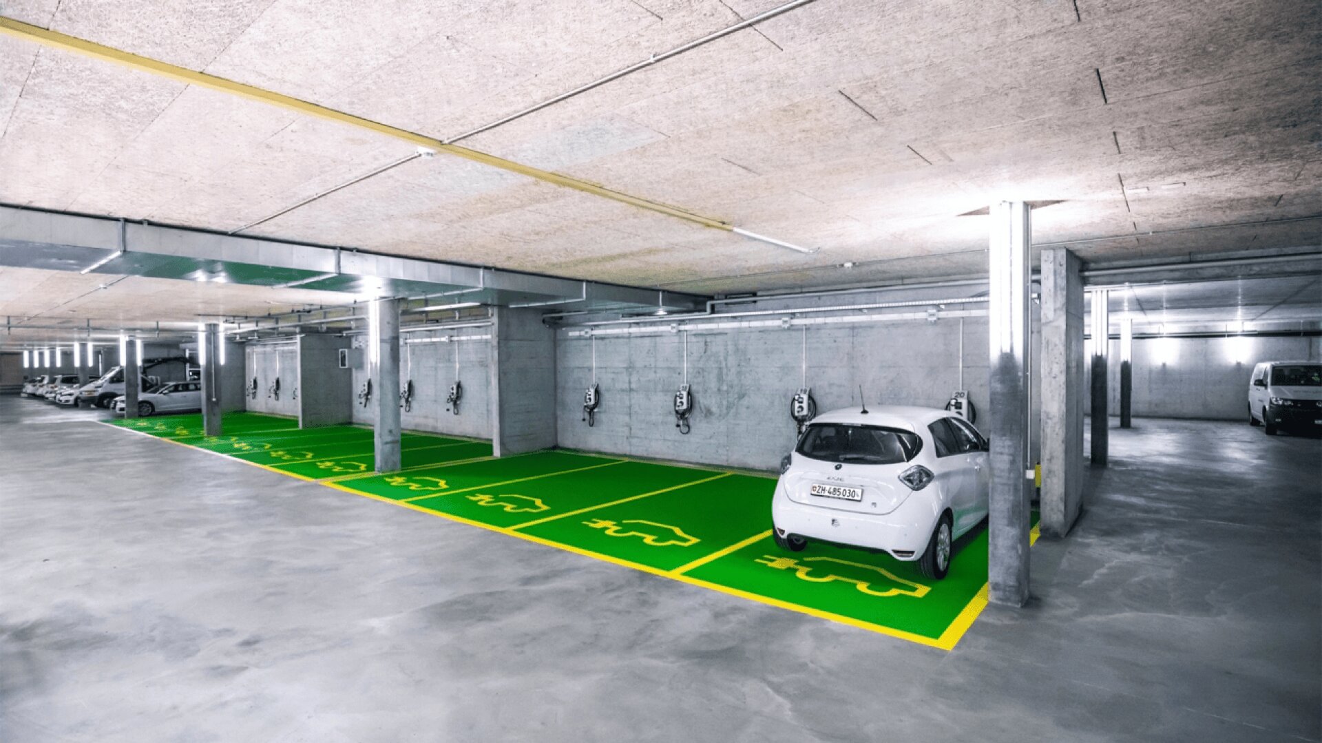 Eine moderne Tiefgarage mit ausgewiesenen grünen Parkplätzen für Elektrofahrzeuge, mit Ladestationen an der Wand; ein weißes Elektroauto ist geparkt und lädt.