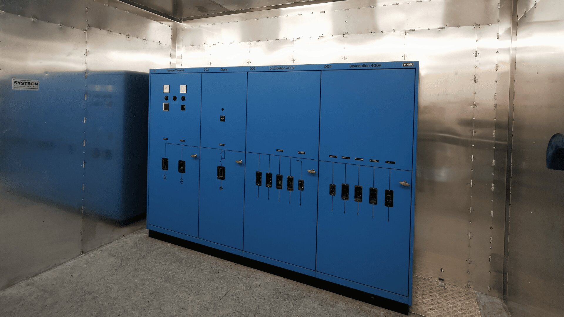 Ein großer blauer Schaltschrank mit mehreren Schaltern, Tasten und Anzeigen ist an einer Metallwand in einem Industrieraum installiert. Ein weiterer blauer Schrank ist auf der linken Seite teilweise sichtbar.