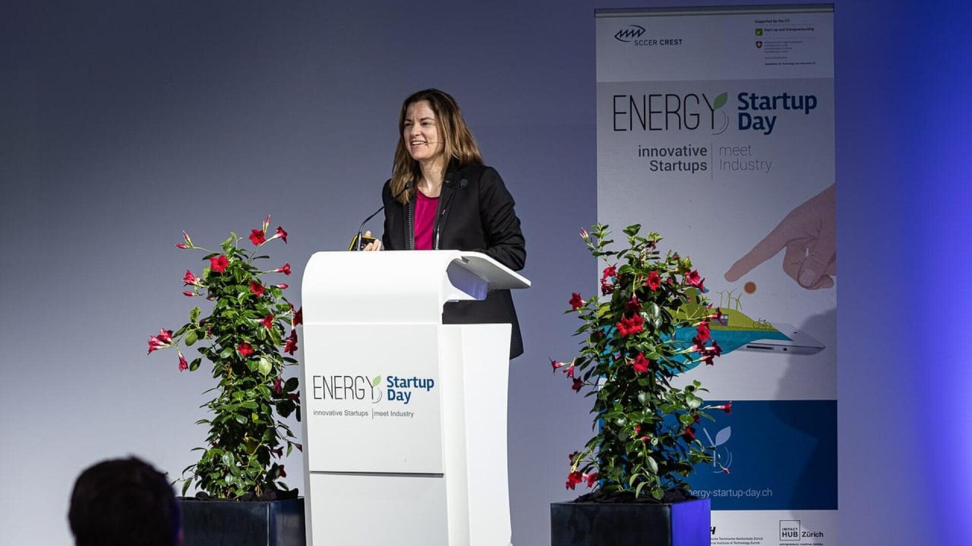 A woman stands at a podium giving a speech at Energy Startup Day. There are two leafy plants flanking the podium and a banner in the background with the events name and graphics about innovation and startups.
