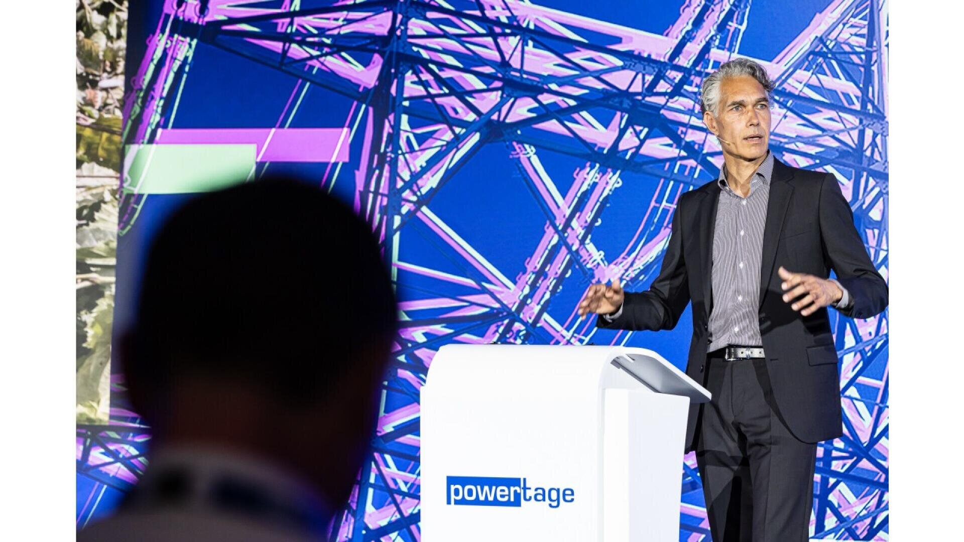 A man in a suit speaks at a podium labeled powertage, with a colorful, abstract background behind him. Another person is visible in the foreground, slightly out of focus.