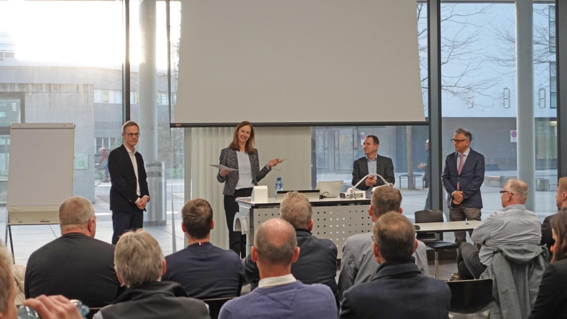 A woman stands and speaks to a seated audience in a conference room, with three men beside her at the front. A blank flip chart, tables, and a large window are visible in the background.