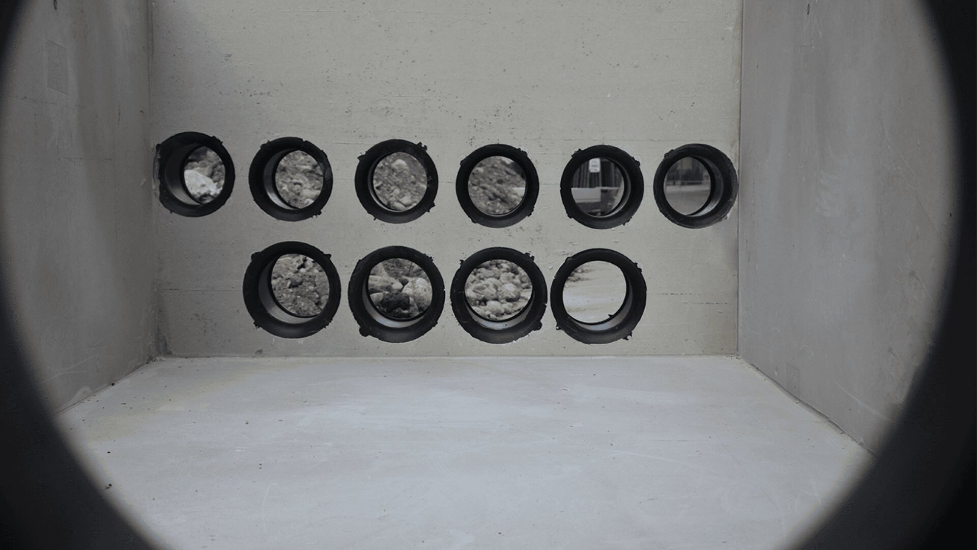A concrete wall with twelve circular openings arranged in two rows. The holes are lined with black plastic and show rocks behind most of them; one opening reveals a blurry scene with colors outside.