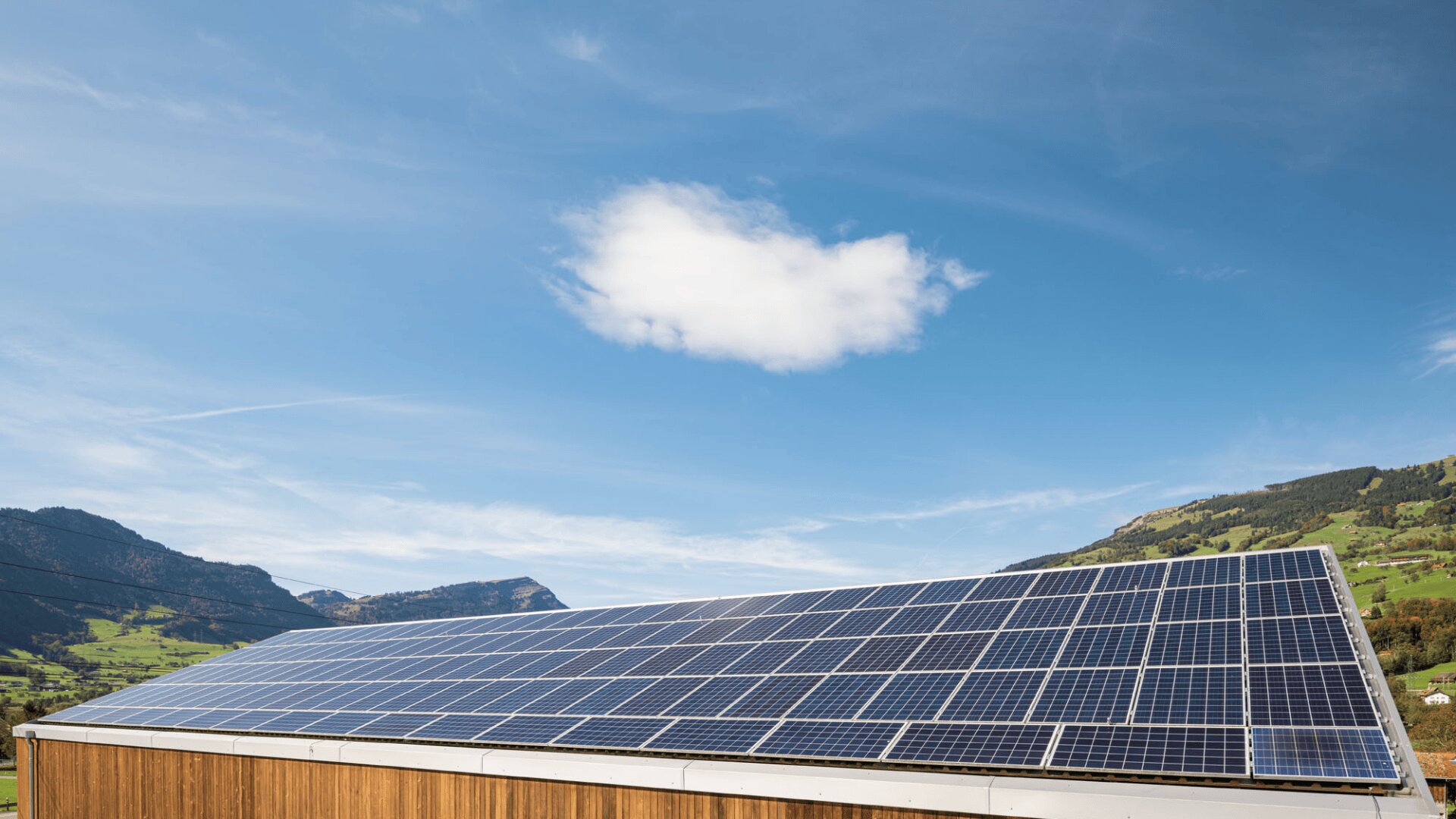 Solar panels installed on a wooden building roof, with a single white cloud in a blue sky and green hills in the background.